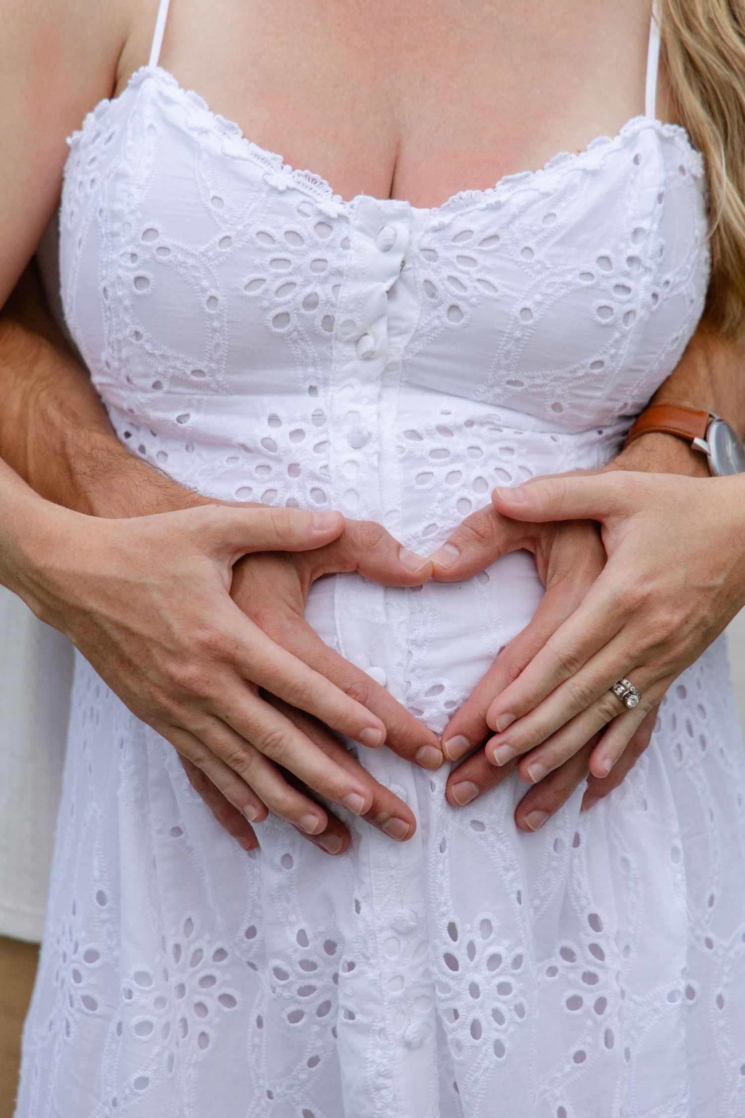Couple forming heart shape with hands on pregnant belly, woman in white eyelet dress wearing engagement ring.