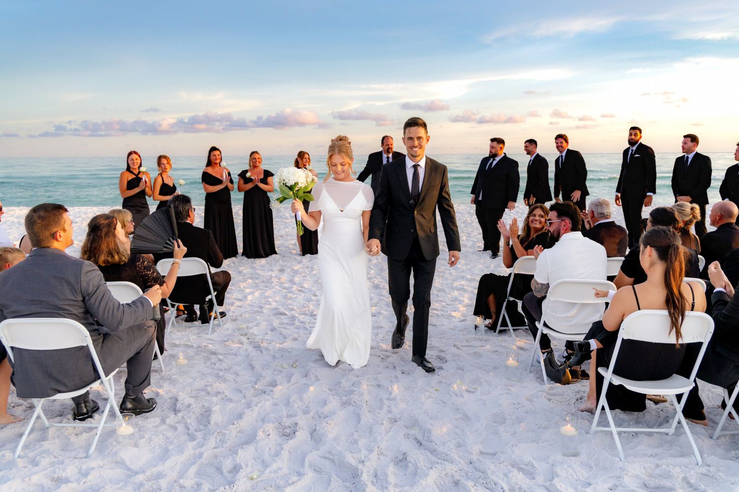 Bride and groom walk down sandy beach aisle at sunset, surrounded by guests in black formal attire.