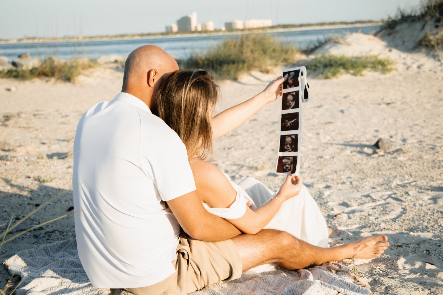 Couple sitting on beach sand looking at ultrasound photos during a maternity photo session.
