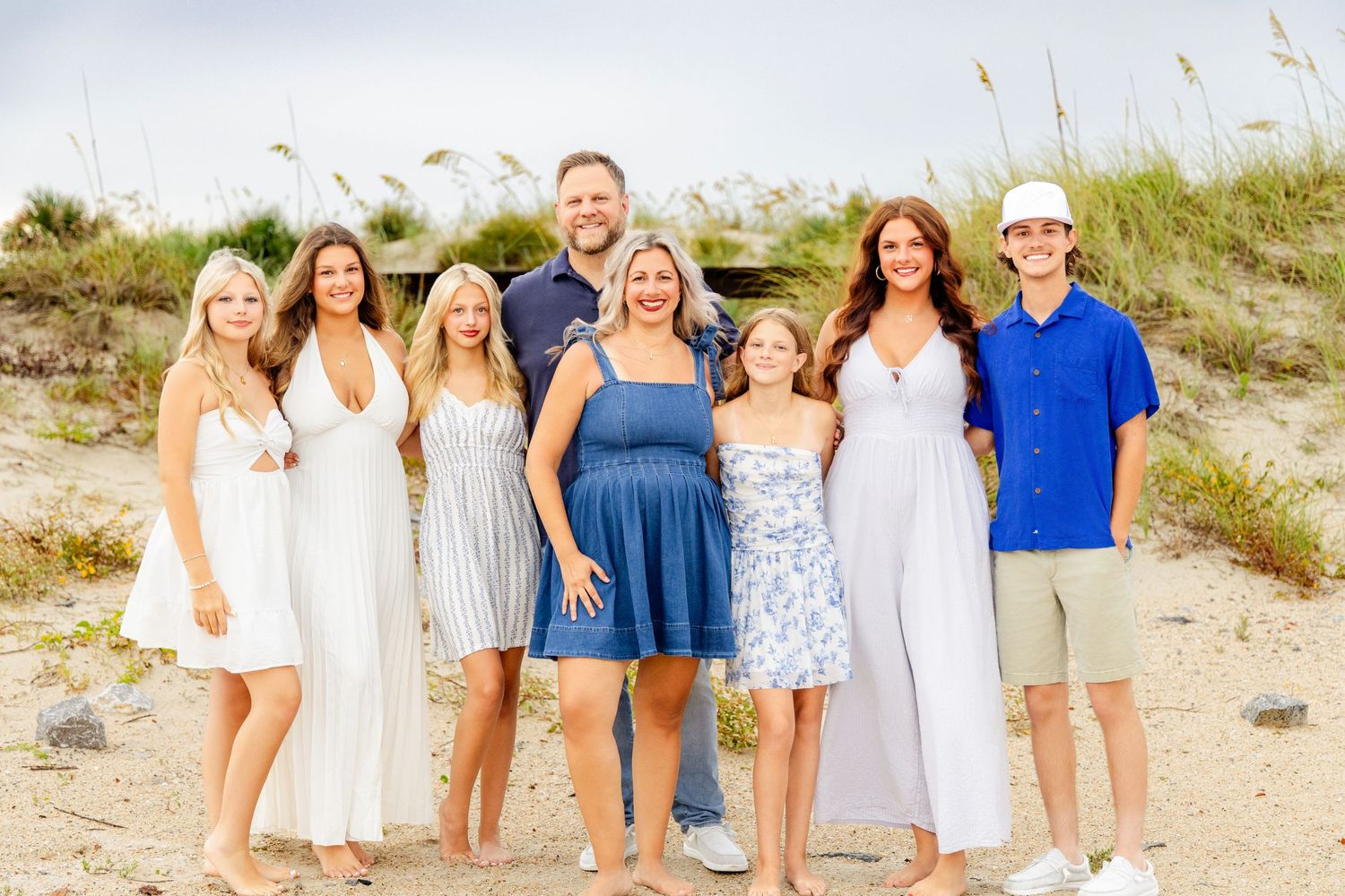 A joyful family of eight poses together on a sandy beach, dressed in coordinating white and blue outfits.