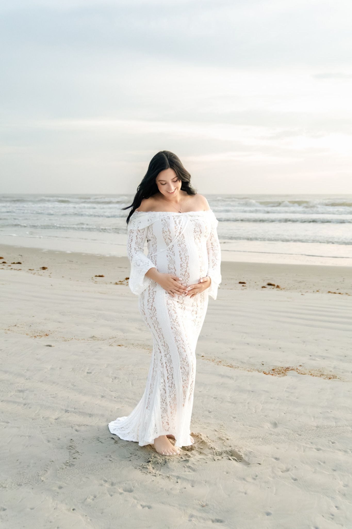 Pregnant woman in white lace off-shoulder gown standing on sandy beach at sunset, cradling baby bump.