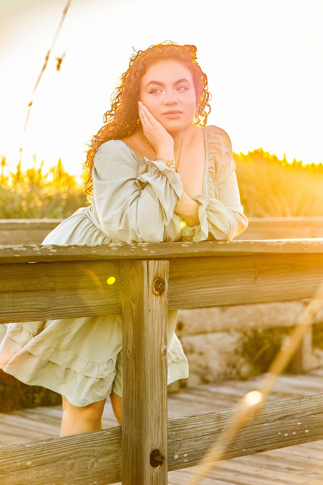 Young woman with curly hair in a sage green dress leaning on a wooden railing at golden hour near beach grasses.