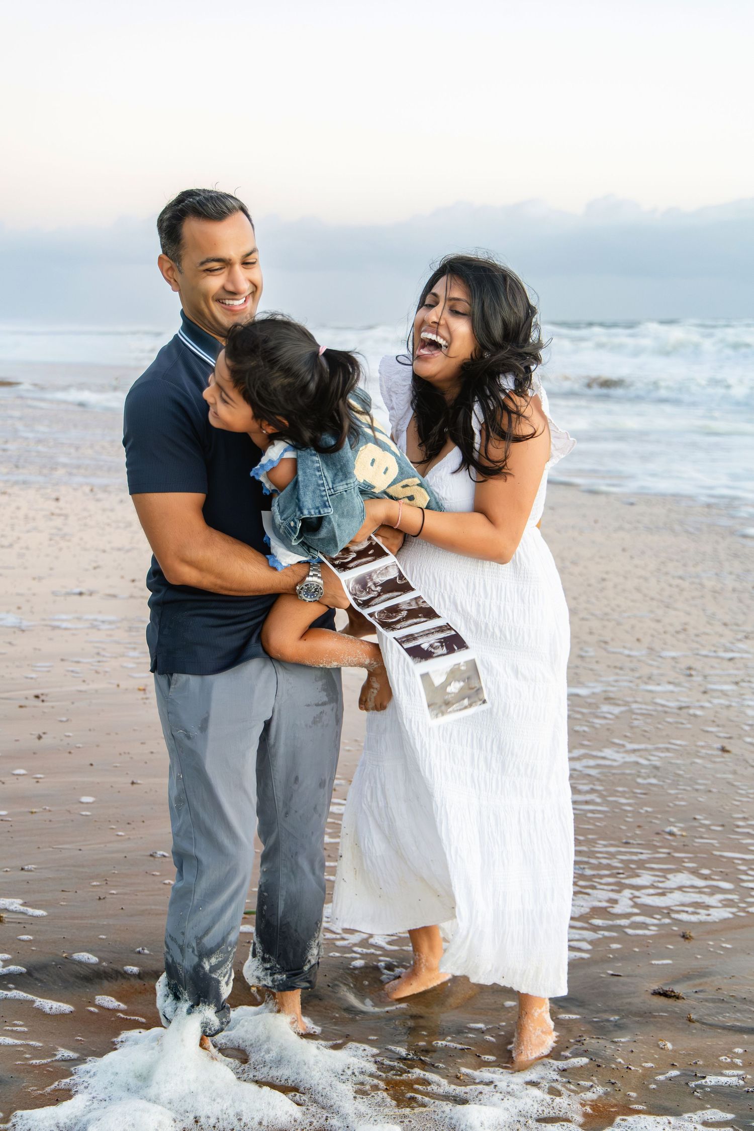 Smiling couple laughing on a beach at sunset, holding their young child and a strip of ultrasound photos in a pregnancy announcement.