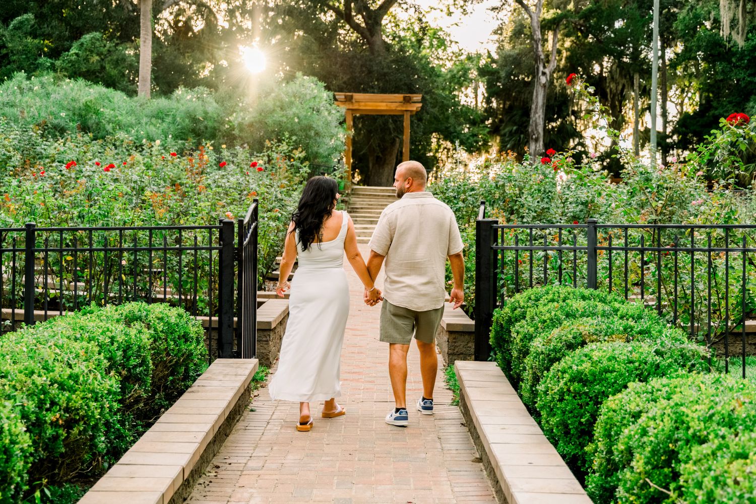 Couple walking hand-in-hand through a lush garden pathway surrounded by roses and greenery at golden hour.