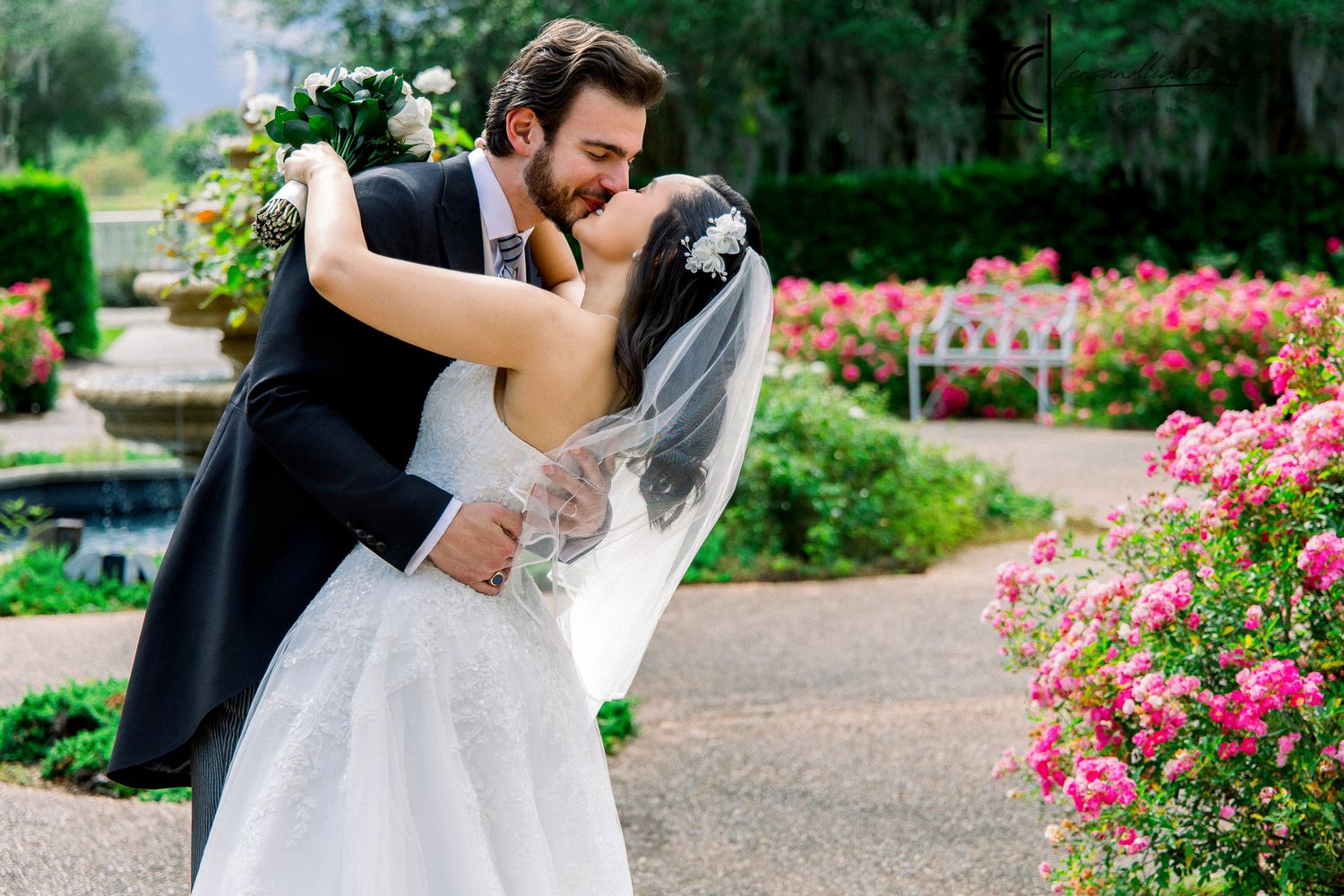 Bride and groom sharing a romantic kiss surrounded by vibrant pink roses in a lush garden setting.