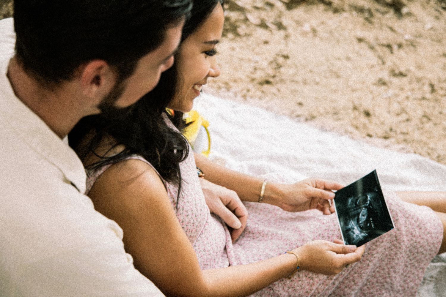 Couple sitting outdoors on a blanket, smiling while looking at an ultrasound photo together.