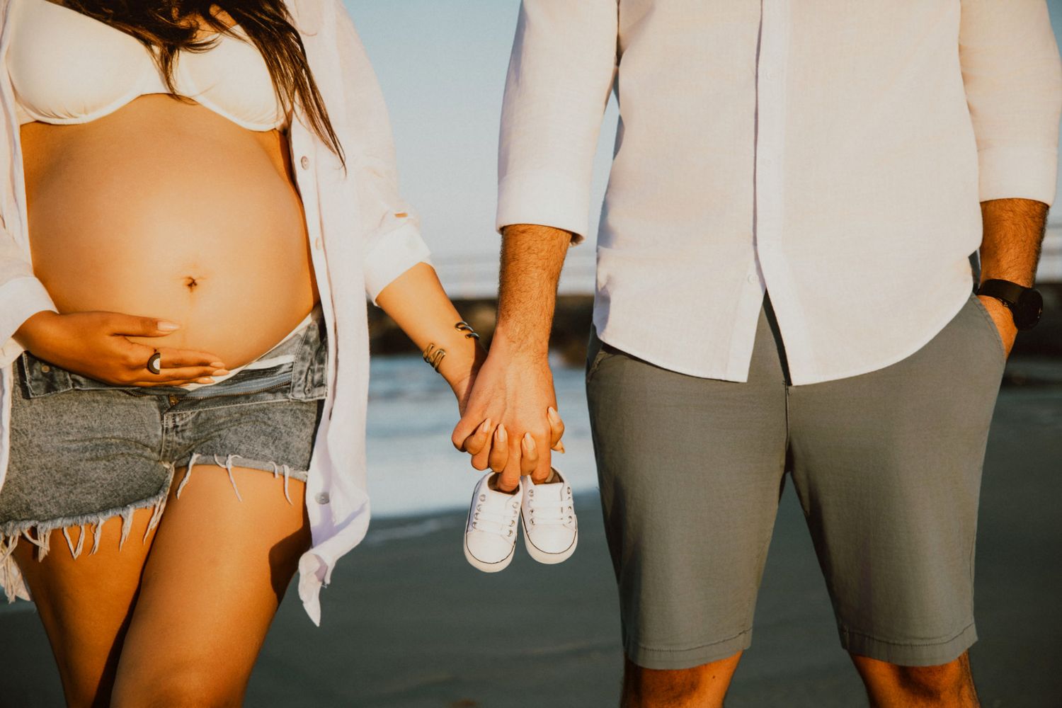 Pregnant couple holding tiny baby shoes and belly outdoors, expecting parents maternity photo at sunset beach.