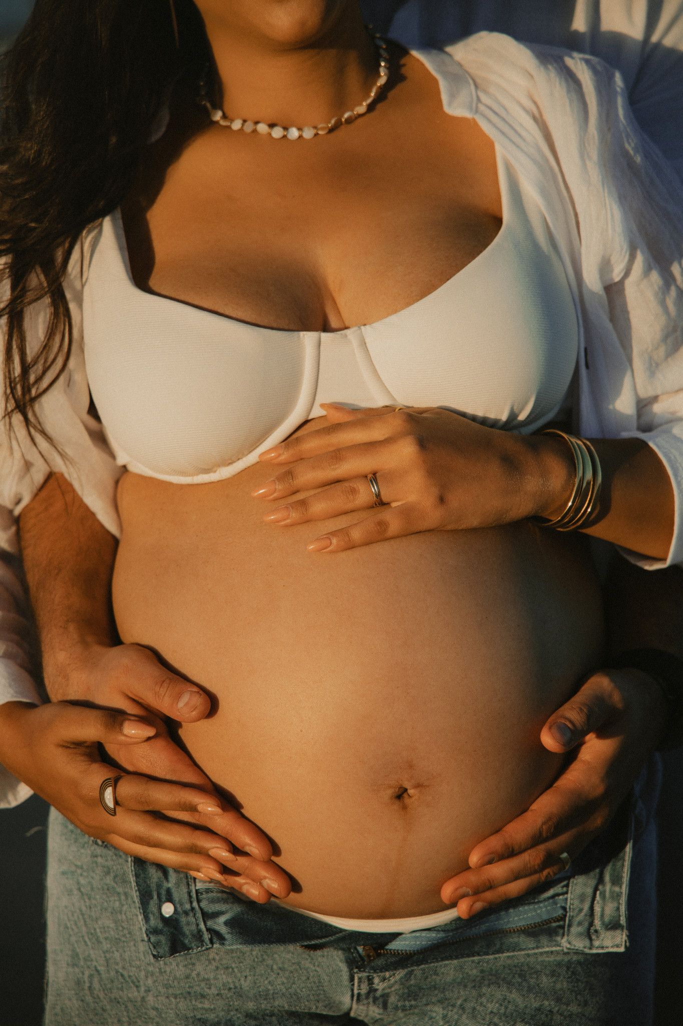 Pregnant woman in white bra and jeans cradling baby bump with partner's hands, golden hour maternity photo.