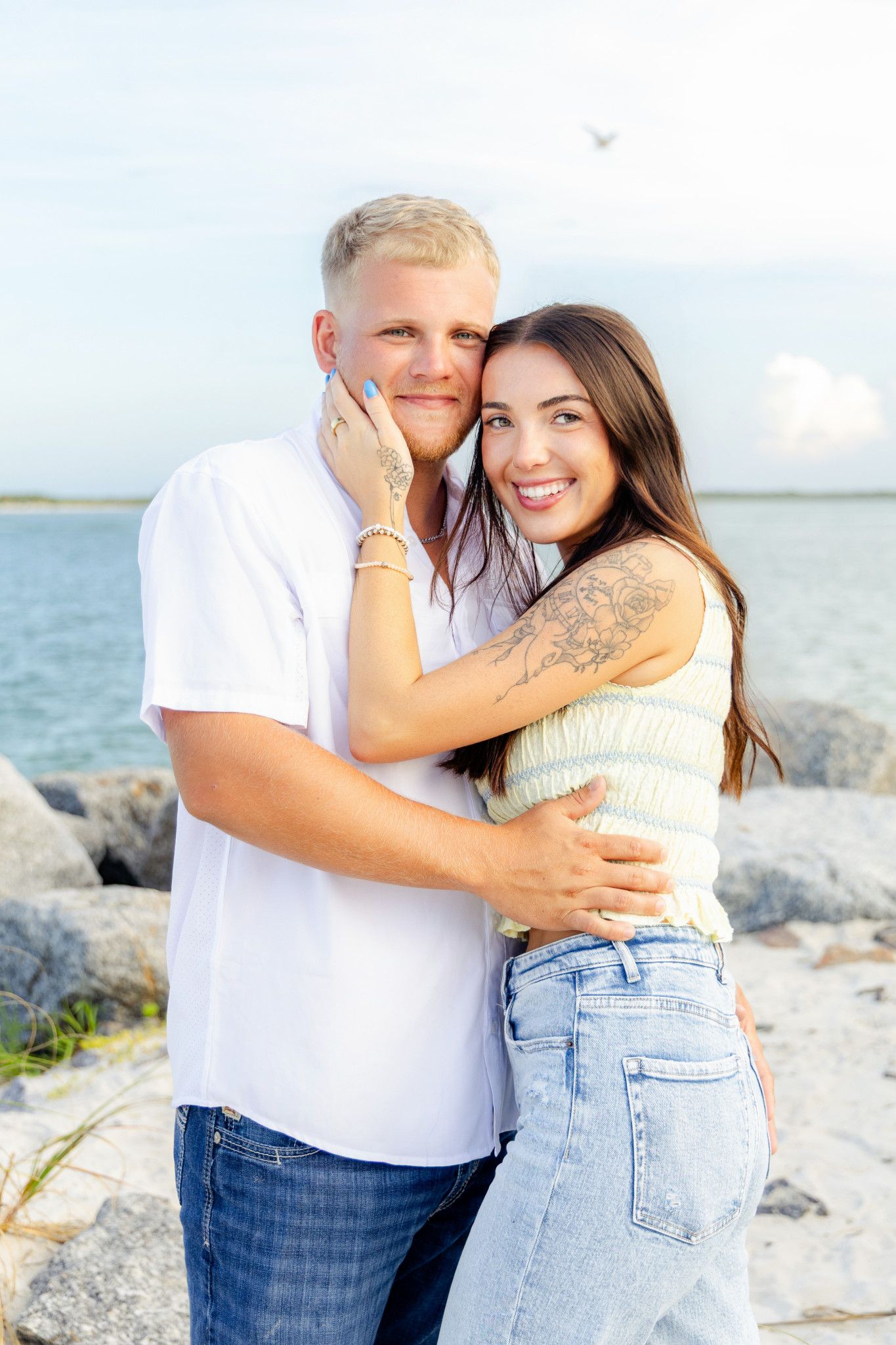 A smiling couple embraces on a rocky beach, woman with tattoos hugging man in white shirt, ocean in background.