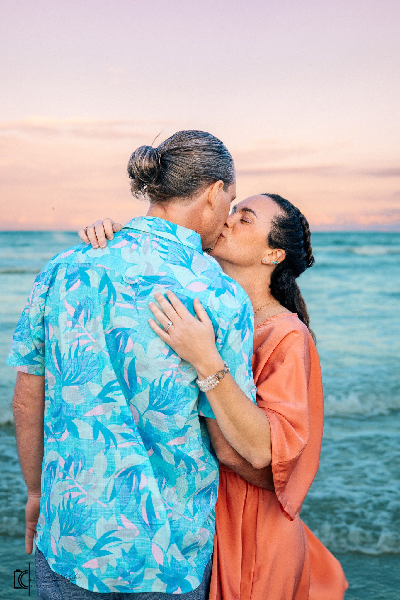A couple shares romantic moments on a beach at sunset wearing coral and blue tropical print outfits.