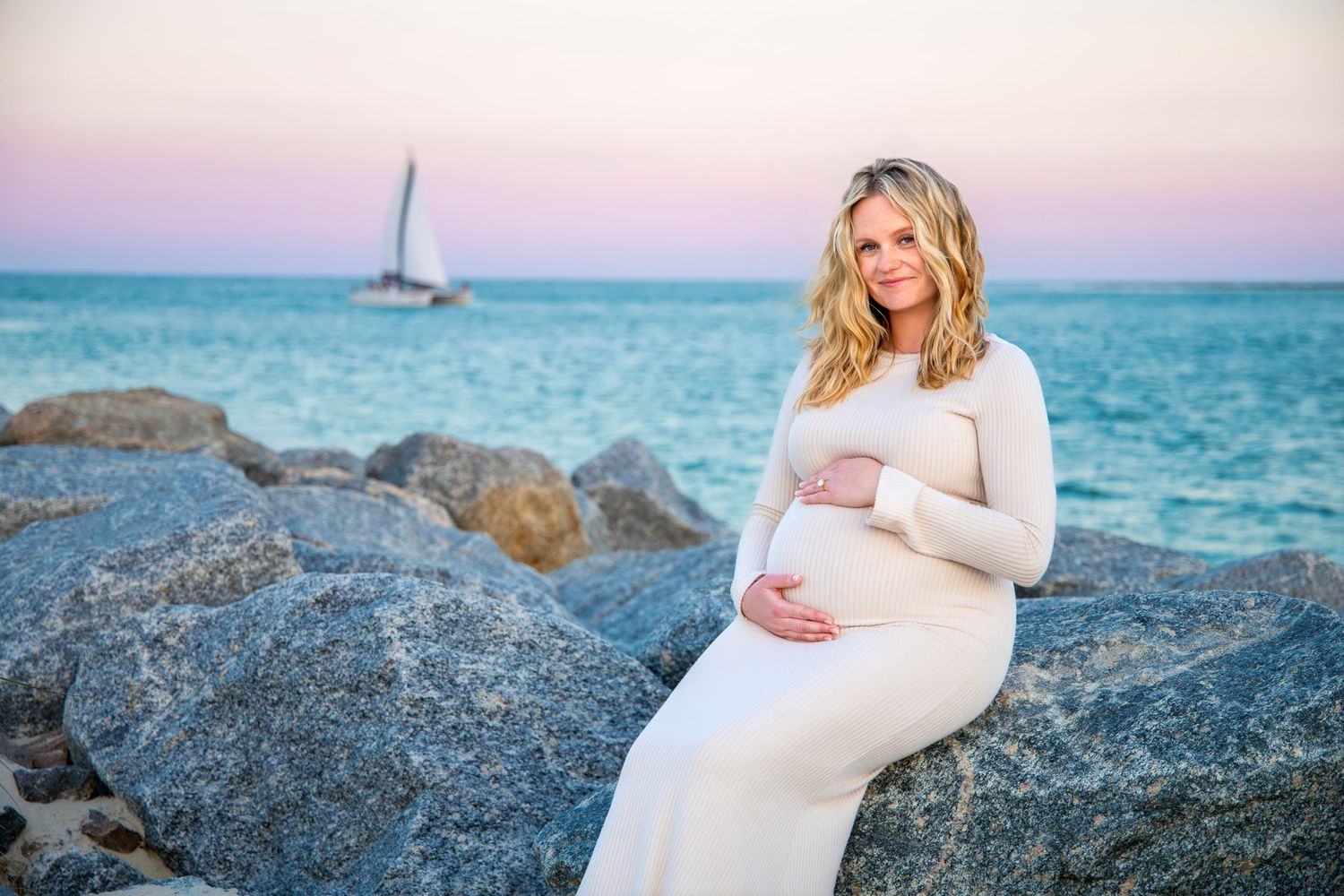 A maternity photoshoot on rocky beach cliffs with turquoise ocean waters and sunset sky in the background.