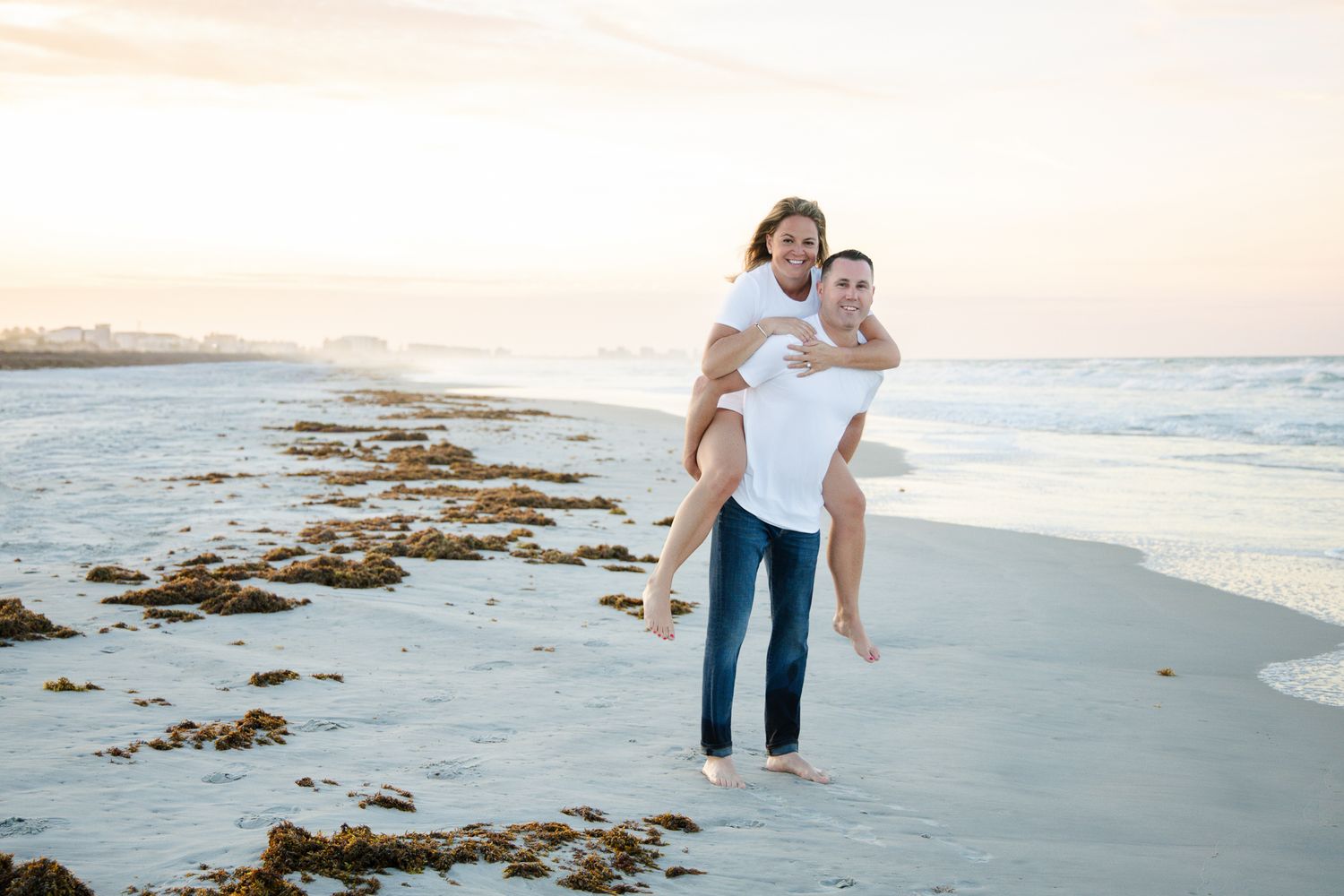 Couple enjoying a piggyback ride on a sandy beach at sunset, wearing casual white tops and jeans.