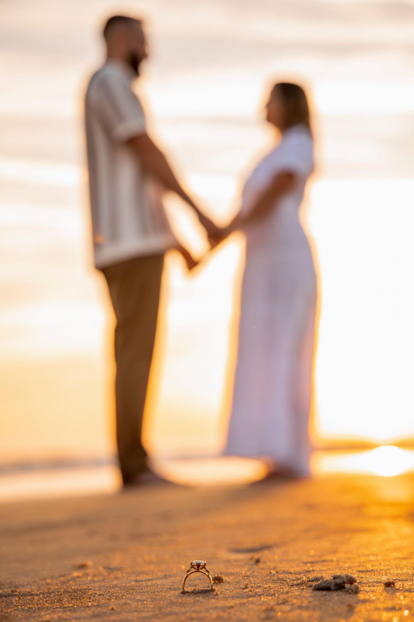 Wedding rings in focus on beach sand with blurred silhouettes of couple holding hands at sunset.