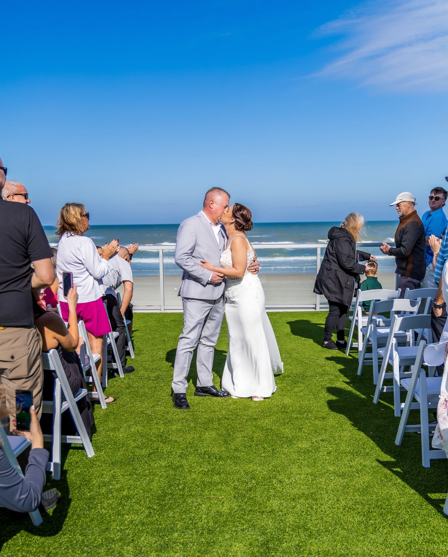 Wedding guests seated on white chairs on a green lawn overlooking a sunny beach with bright blue ocean views.