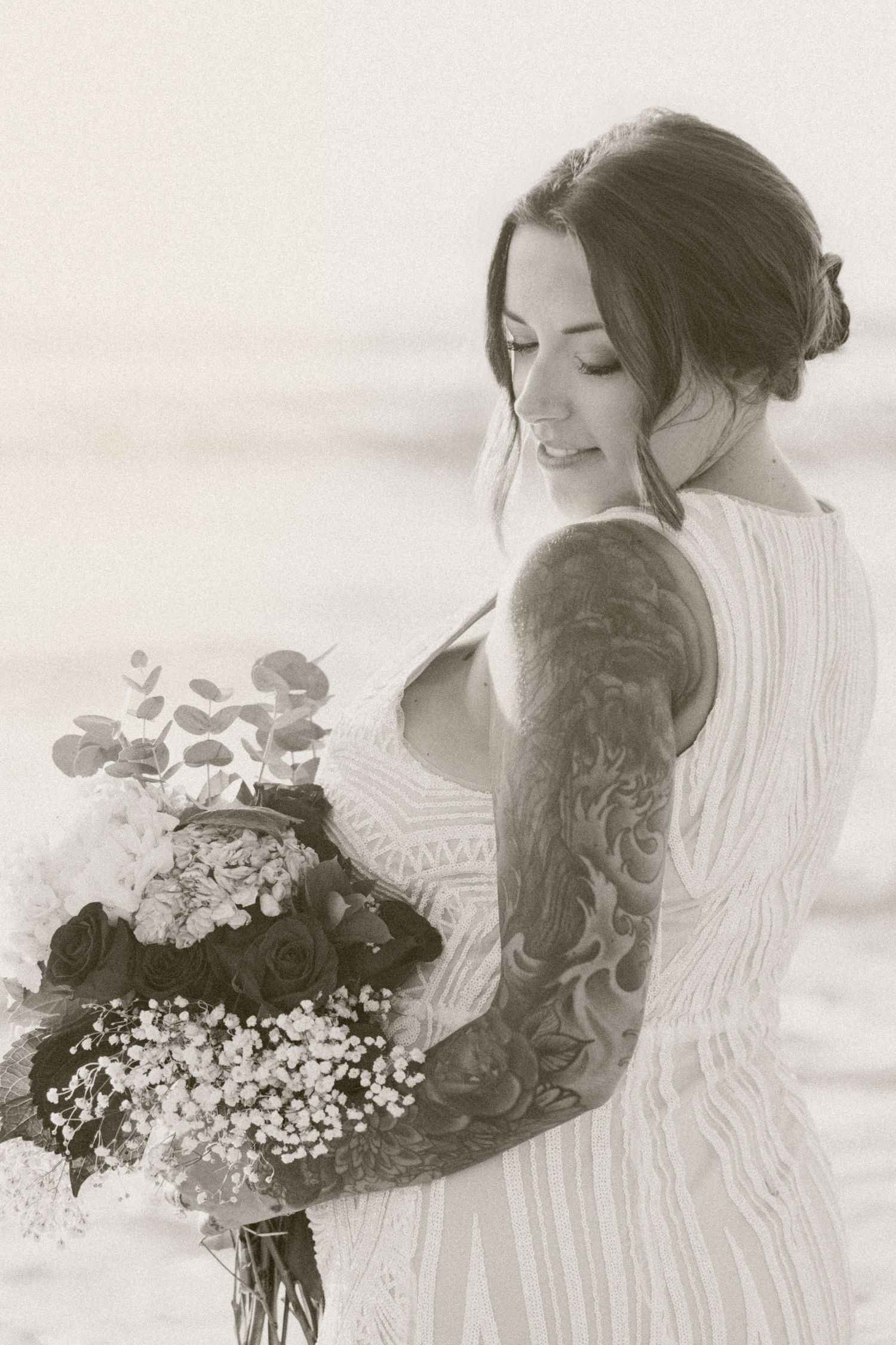 Black and white photo of tattooed bride in lace dress holding bouquet of roses and wildflowers on beach.