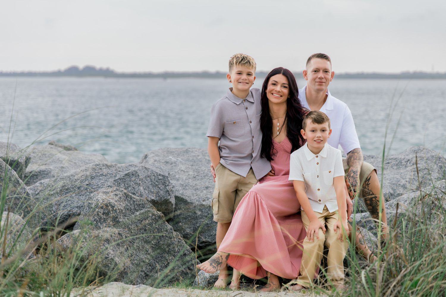 Family of four sitting on rocky shoreline, smiling together with calm water and greenery in the background.