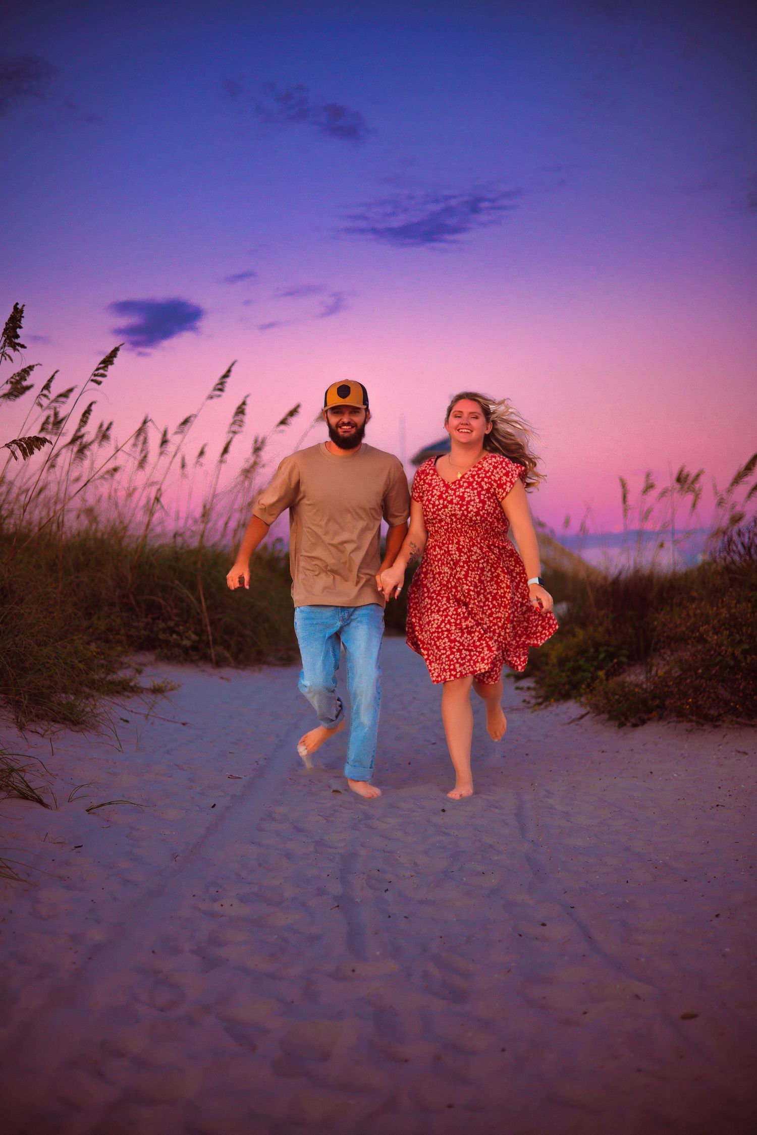 Couple walking together on sandy beach dunes at sunset with tall grass silhouettes against purple sky.