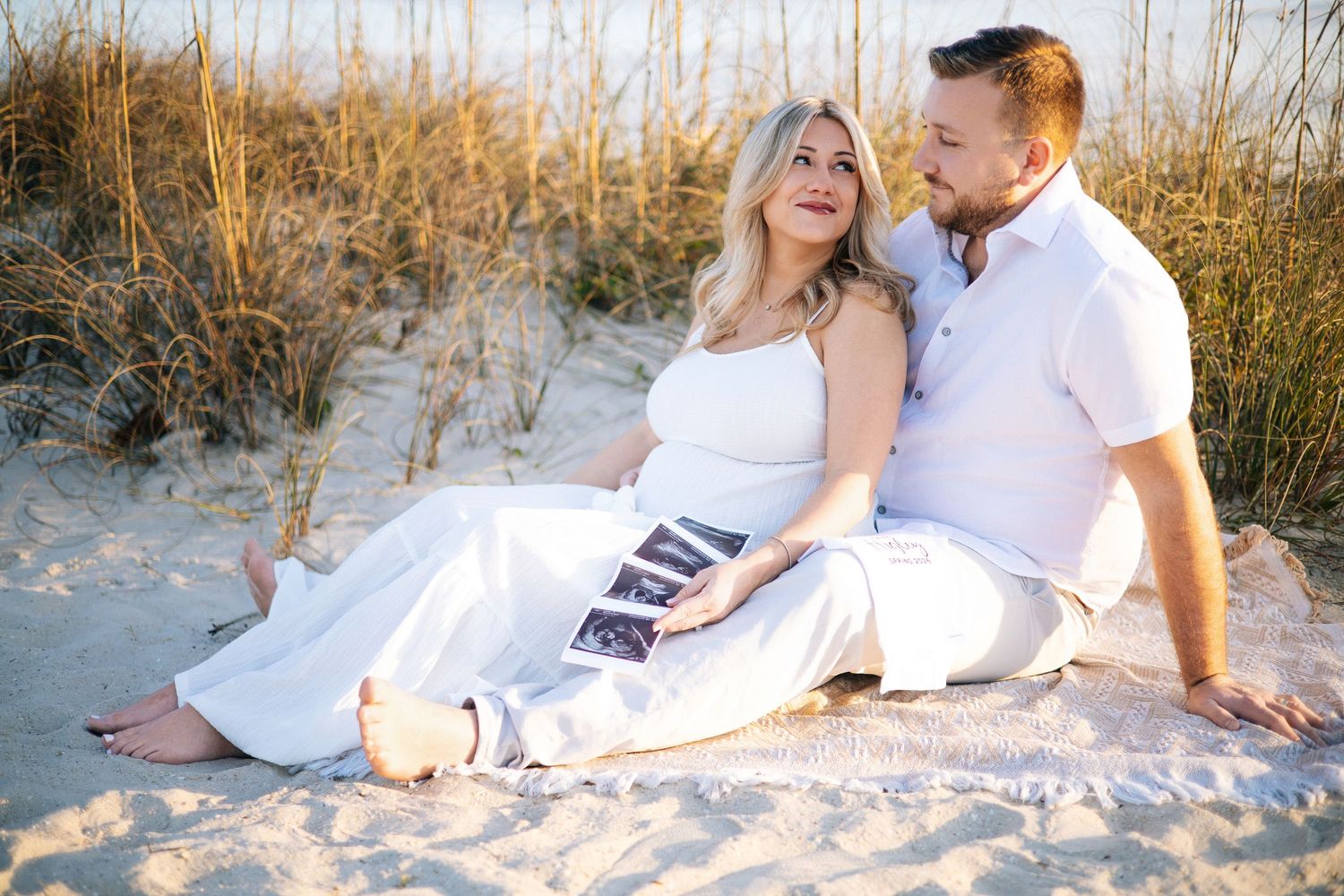 Romantic couple in white outfits sitting together on sandy beach during golden hour sunset.