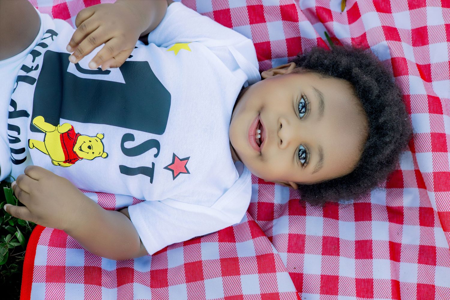 Cute toddler with curly hair lying on a red checkered blanket wearing a Winnie the Pooh shirt.
