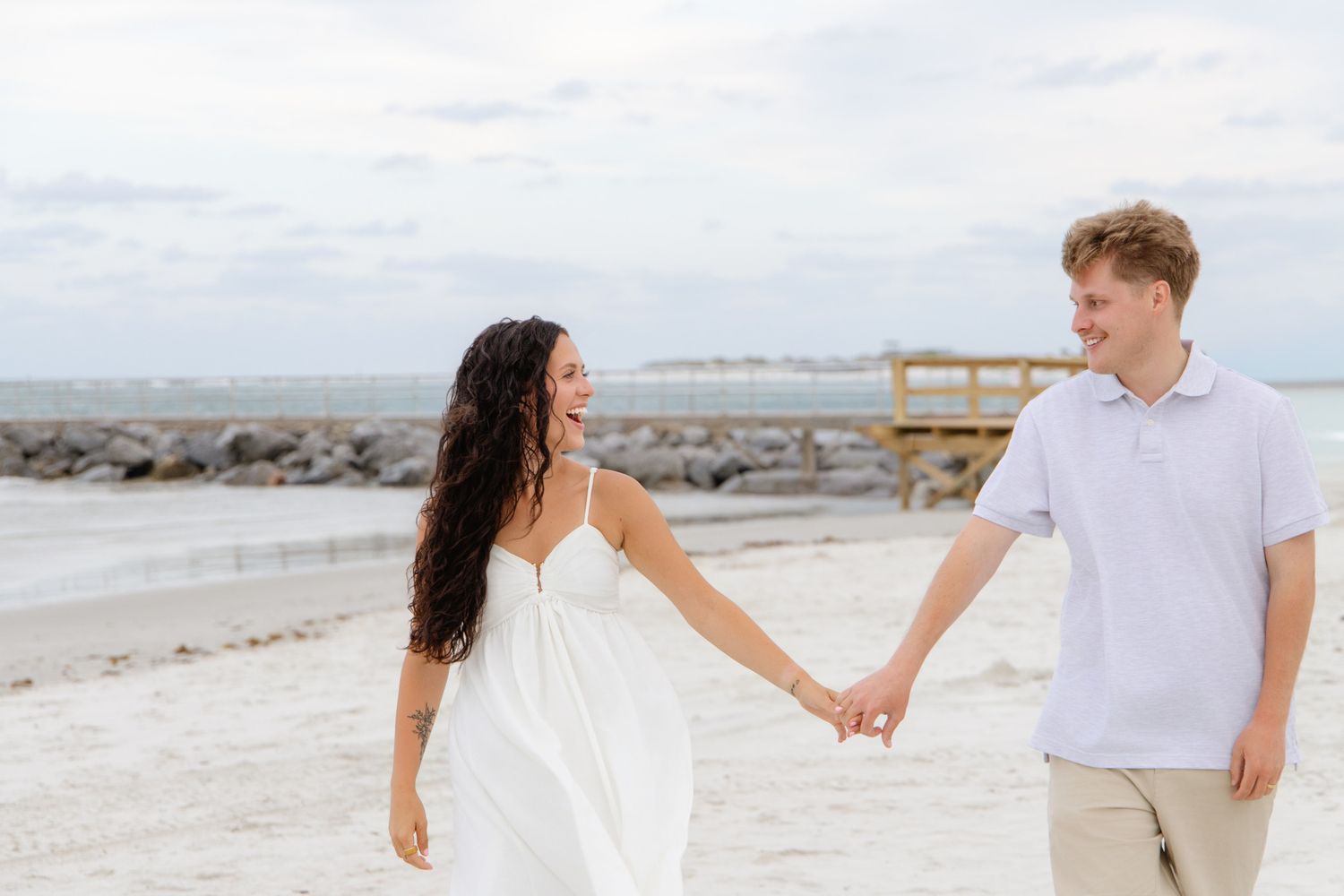Couple holding hands and smiling on a sandy beach, woman in white dress, man in light blue polo shirt.