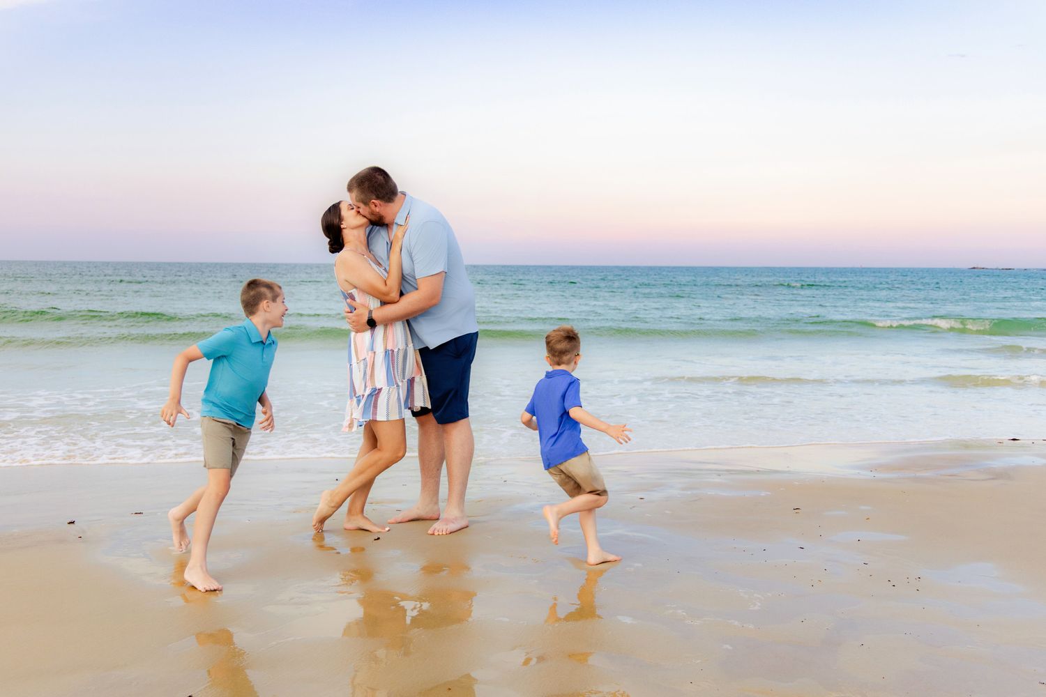 Family with two boys playing on the beach shore at sunset, parents kissing while children run.