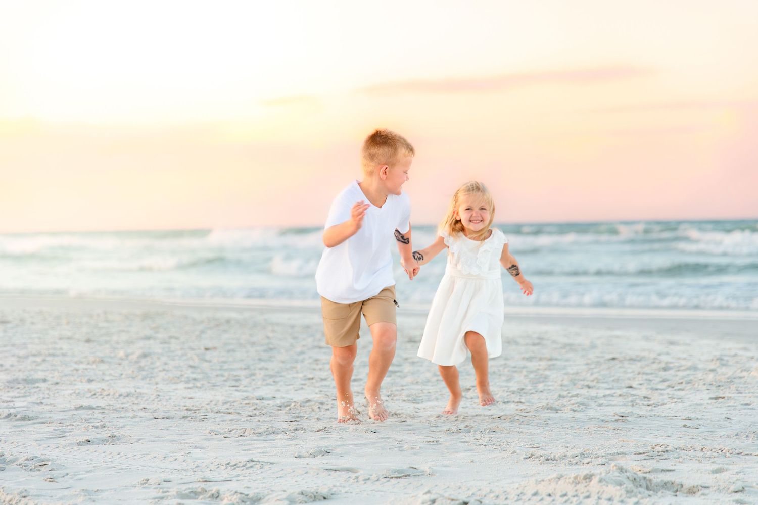 Two young children running joyfully on a sandy beach at sunset, boy in white shirt and girl in white dress.