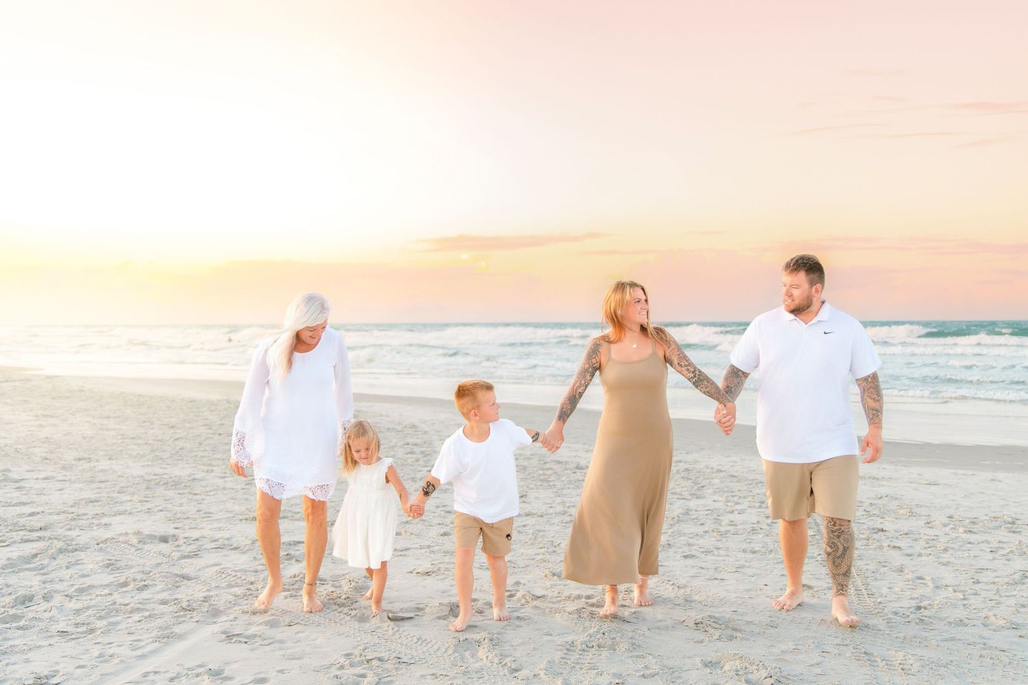 Family of five walking hand-in-hand on a sandy beach at sunset, wearing white and neutral tones.