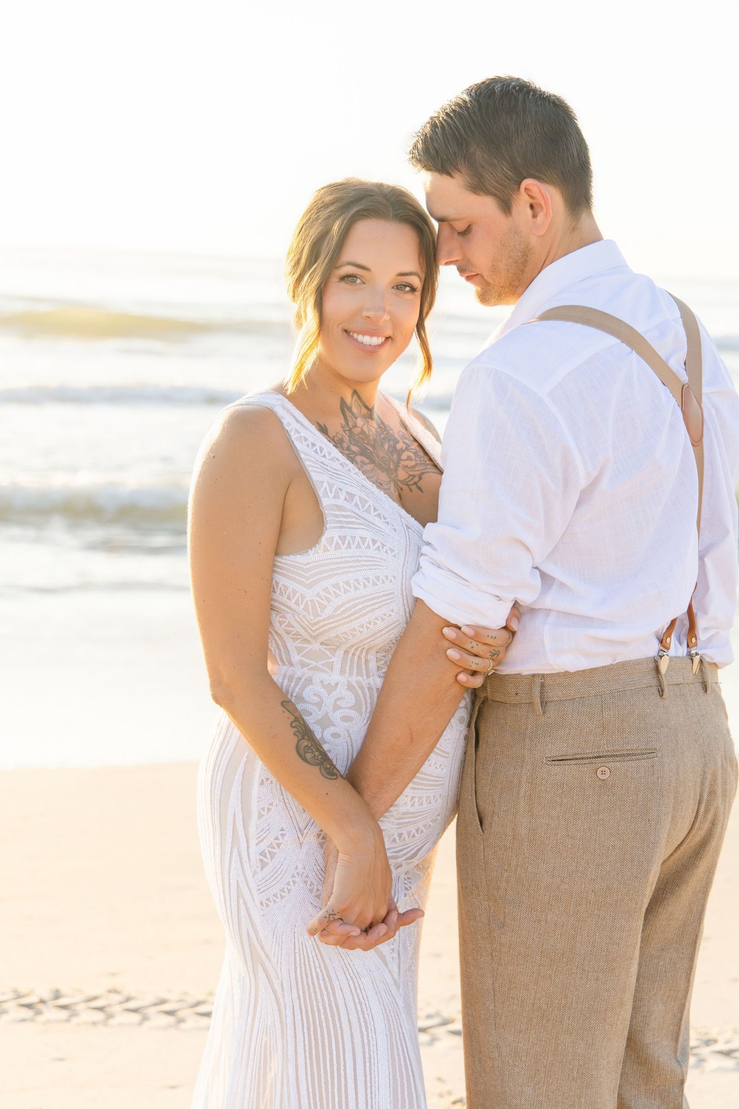 Bride and groom embrace on a sunlit beach, she smiles in a lace gown as he looks down tenderly.