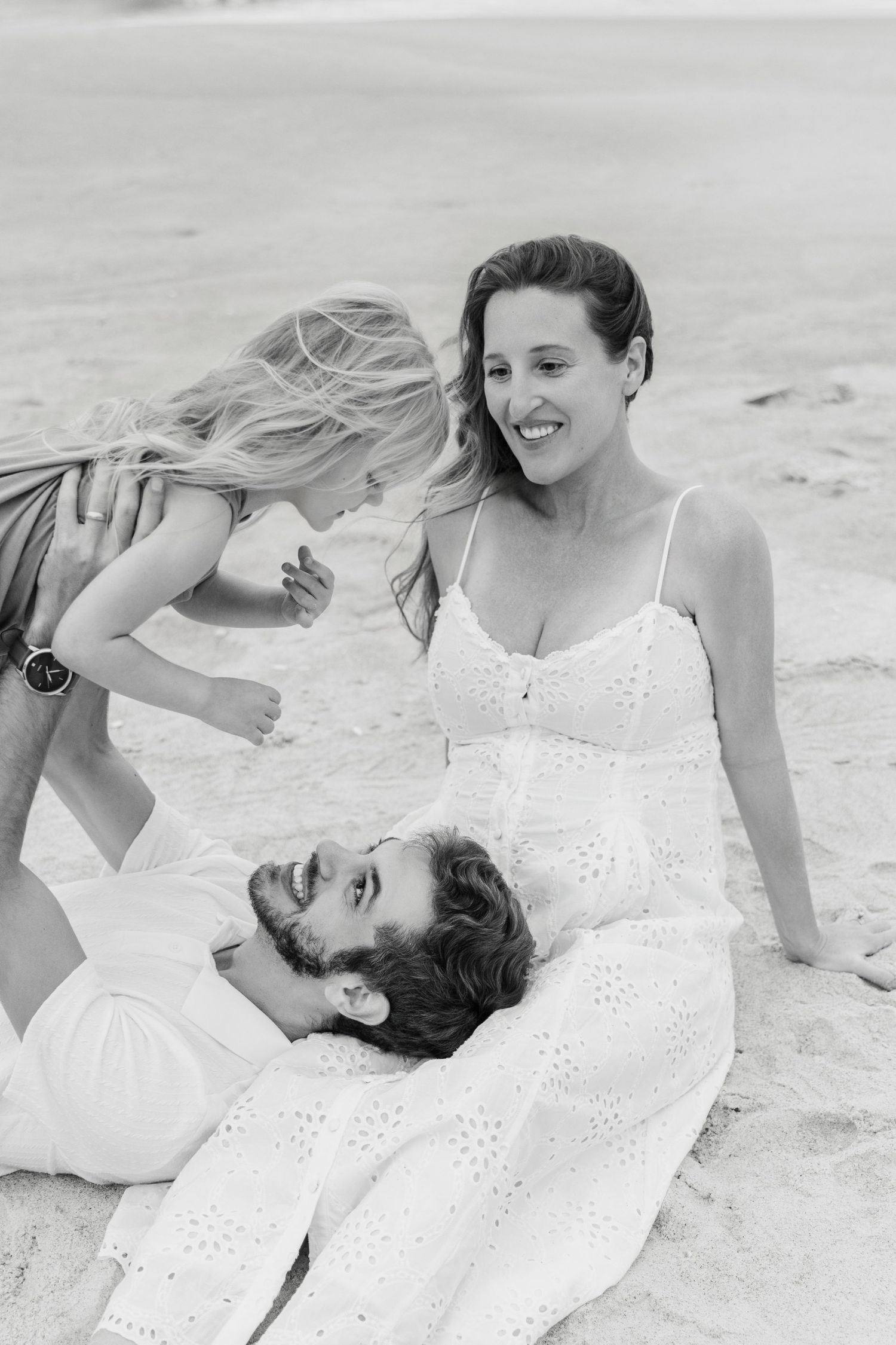 Black and white beach photo of a smiling woman in white dress with bearded man lying in her lap and blonde child.