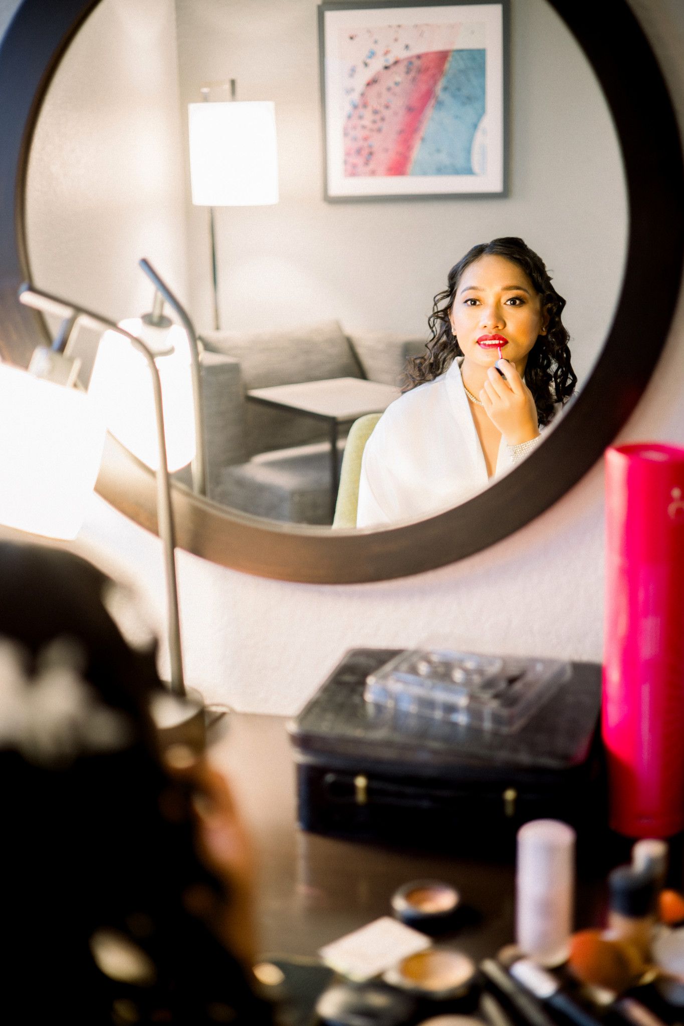 Bride in white robe applying lipstick at vanity mirror surrounded by makeup products.
