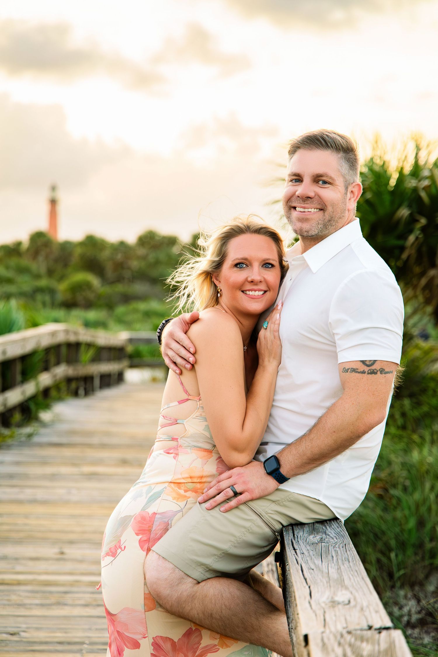 Couple embracing on a wooden boardwalk near a lighthouse at sunset, woman in floral dress, man in white shirt.