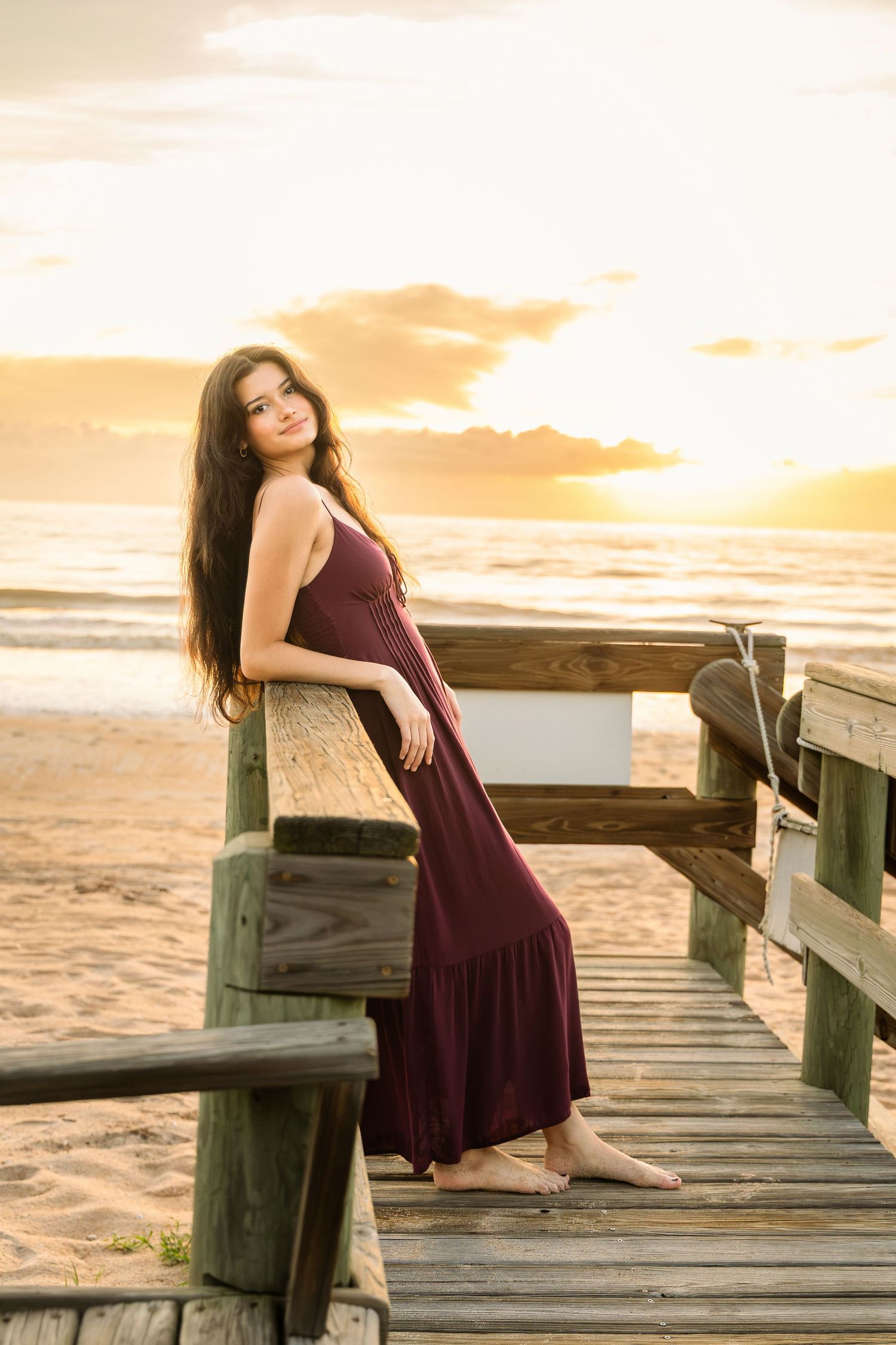 A young woman in a burgundy maxi dress poses on a wooden boardwalk at sunset by the beach.