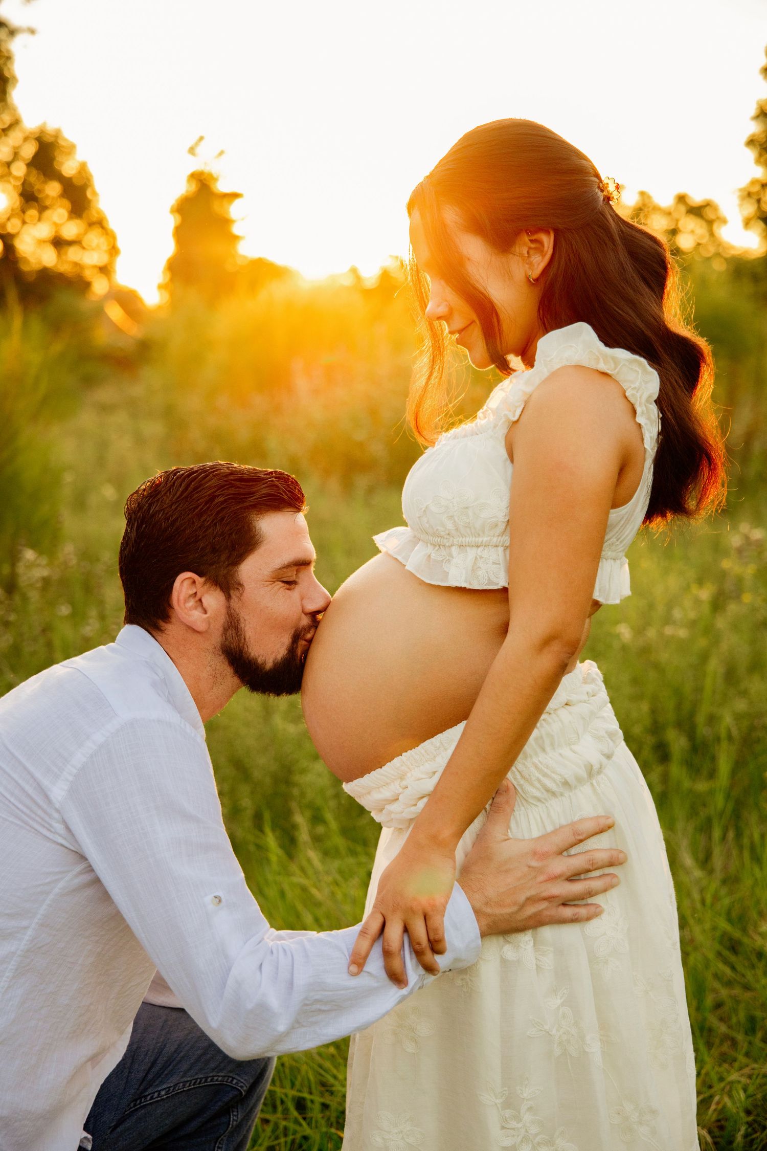 A man kneels kissing a pregnant woman's belly in a sunlit field during golden hour maternity photoshoot.