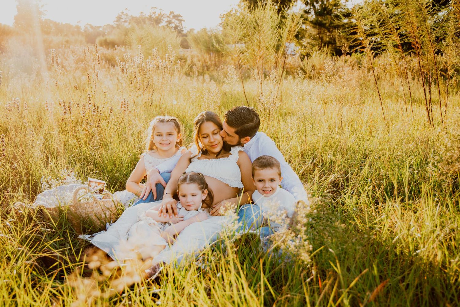 Family maternity photo session in golden field, parents and three children sitting together in tall grass at sunset.