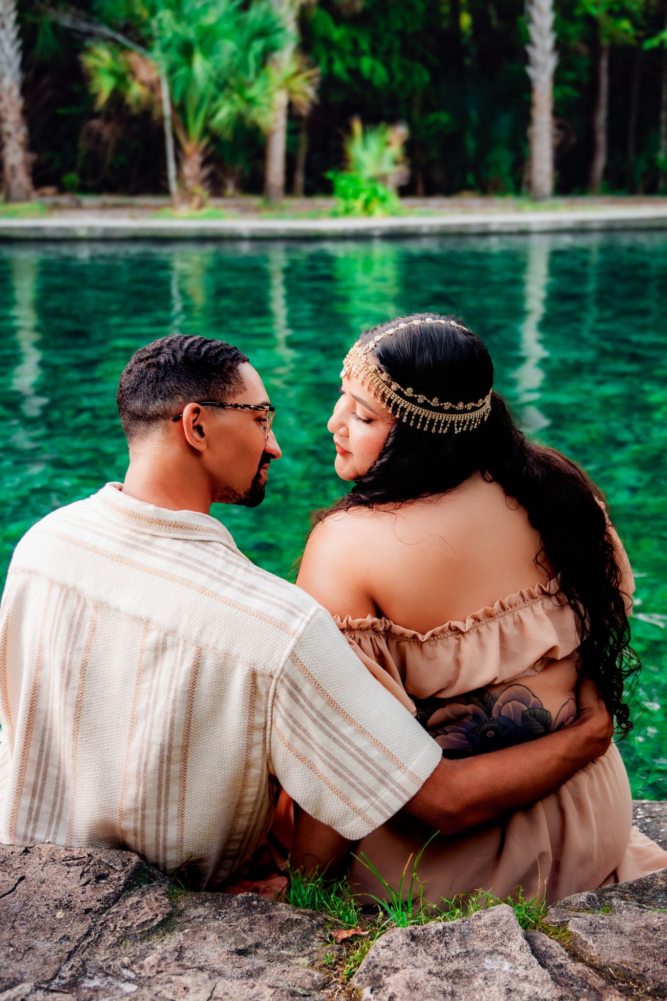 Couple sitting by a turquoise cenote pool, embracing romantically with lush tropical greenery in the background.