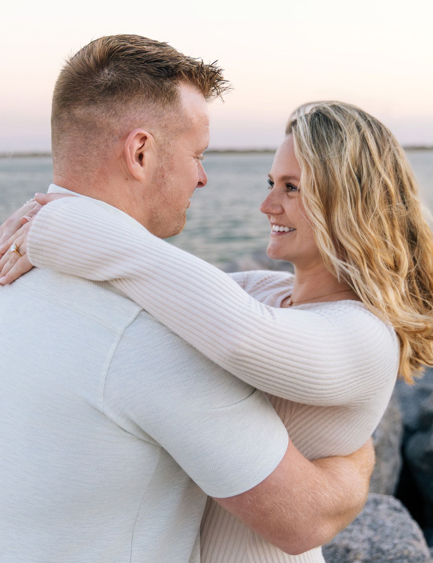 A couple embraces on a beach at sunset wearing white sweaters as they share an intimate moment together.