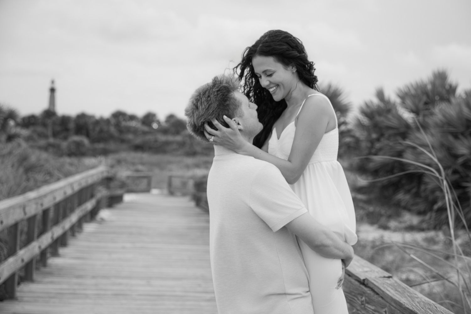 Black and white photo of a couple embracing on a wooden boardwalk near a beach lighthouse.
