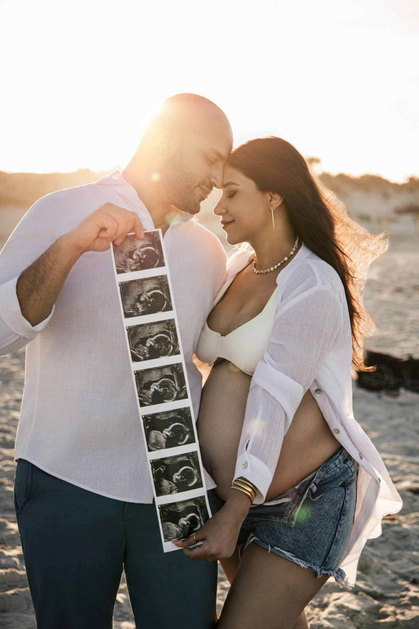 Couple sharing a tender moment on a beach at sunset, holding ultrasound photos while revealing a pregnancy.