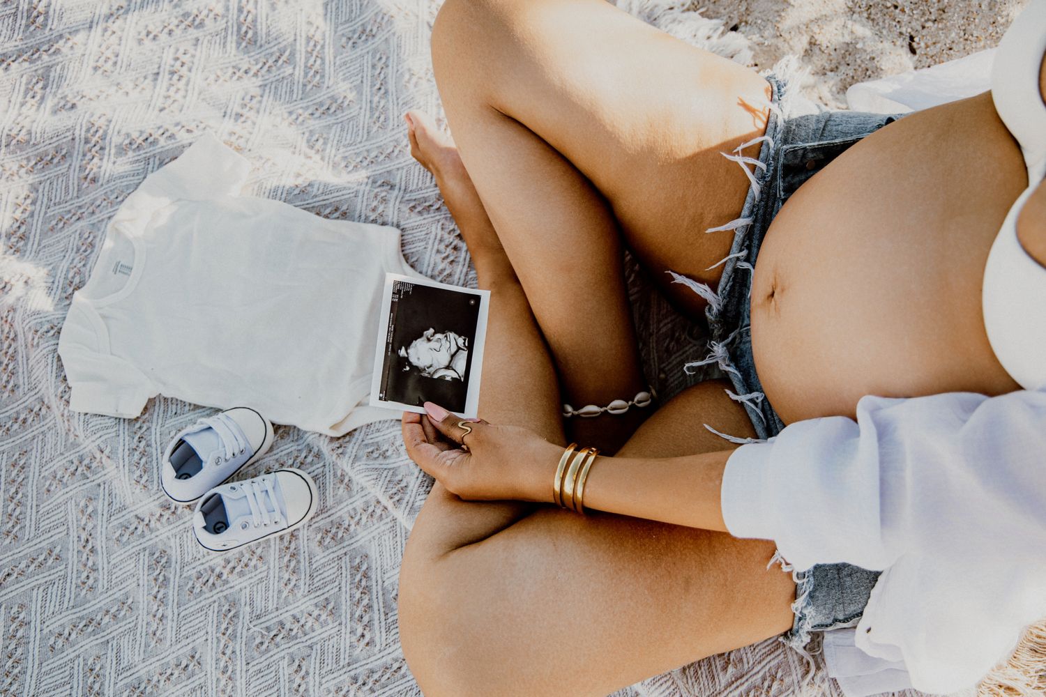 Pregnant woman sitting on blanket holding ultrasound photo with baby shoes and clothing nearby.