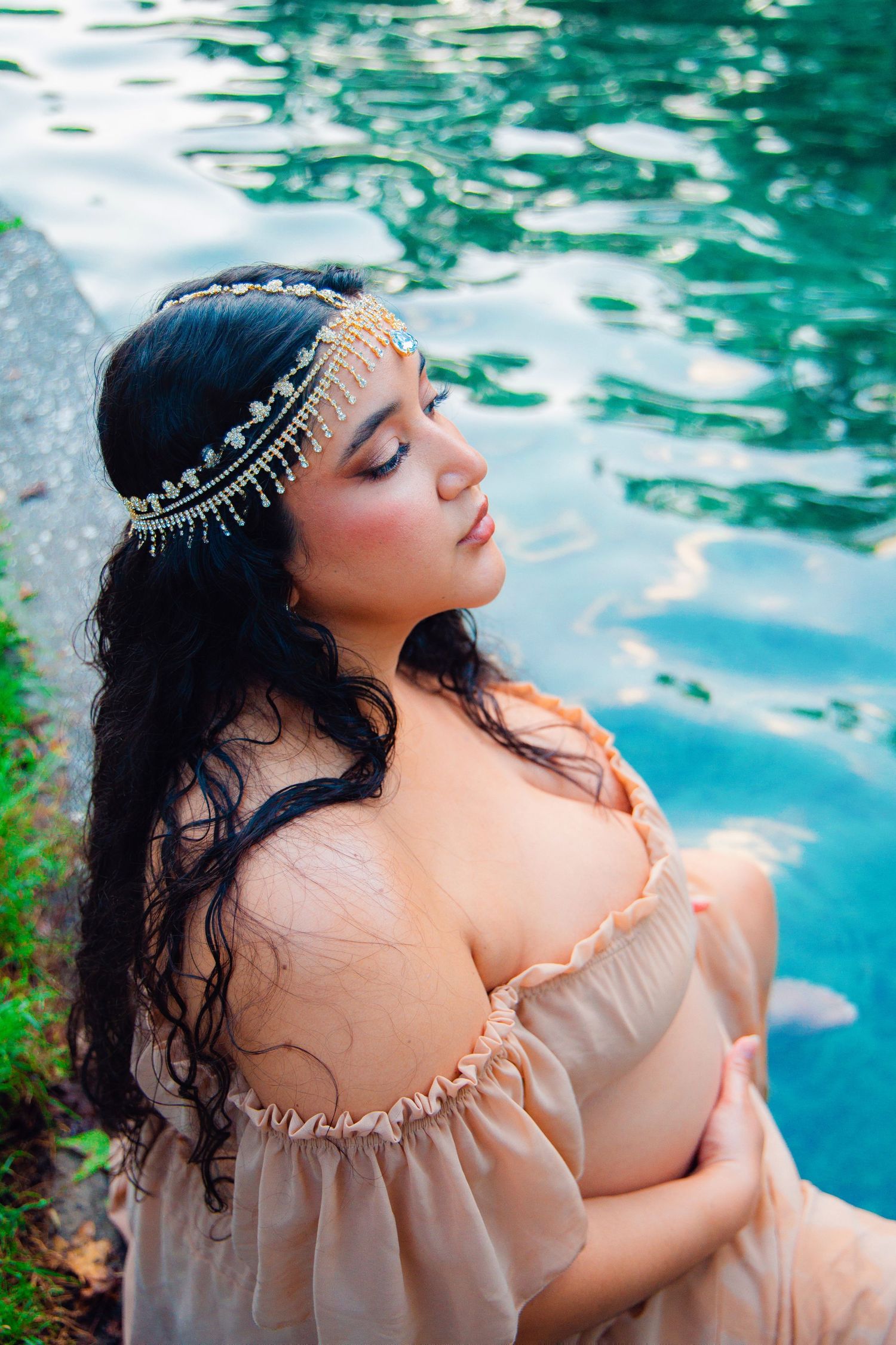 Woman in beige off-shoulder dress with gold headpiece sits peacefully by turquoise water with eyes closed.