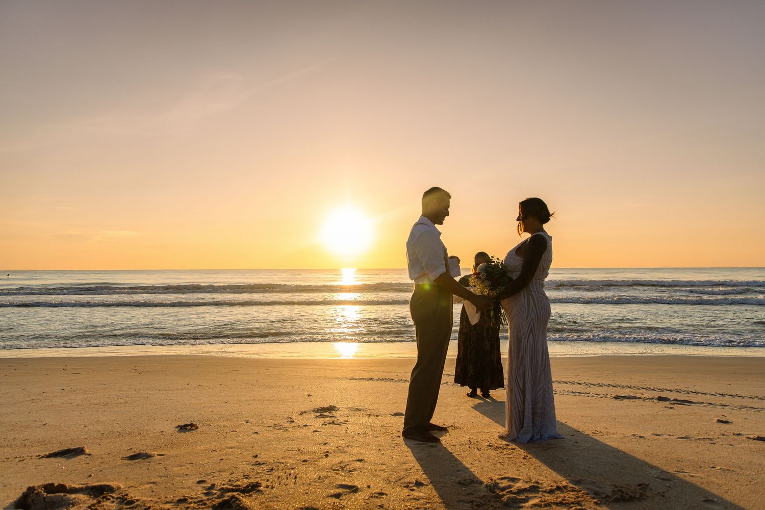 Couple exchanging vows on a sandy beach at golden sunset with waves in the background.