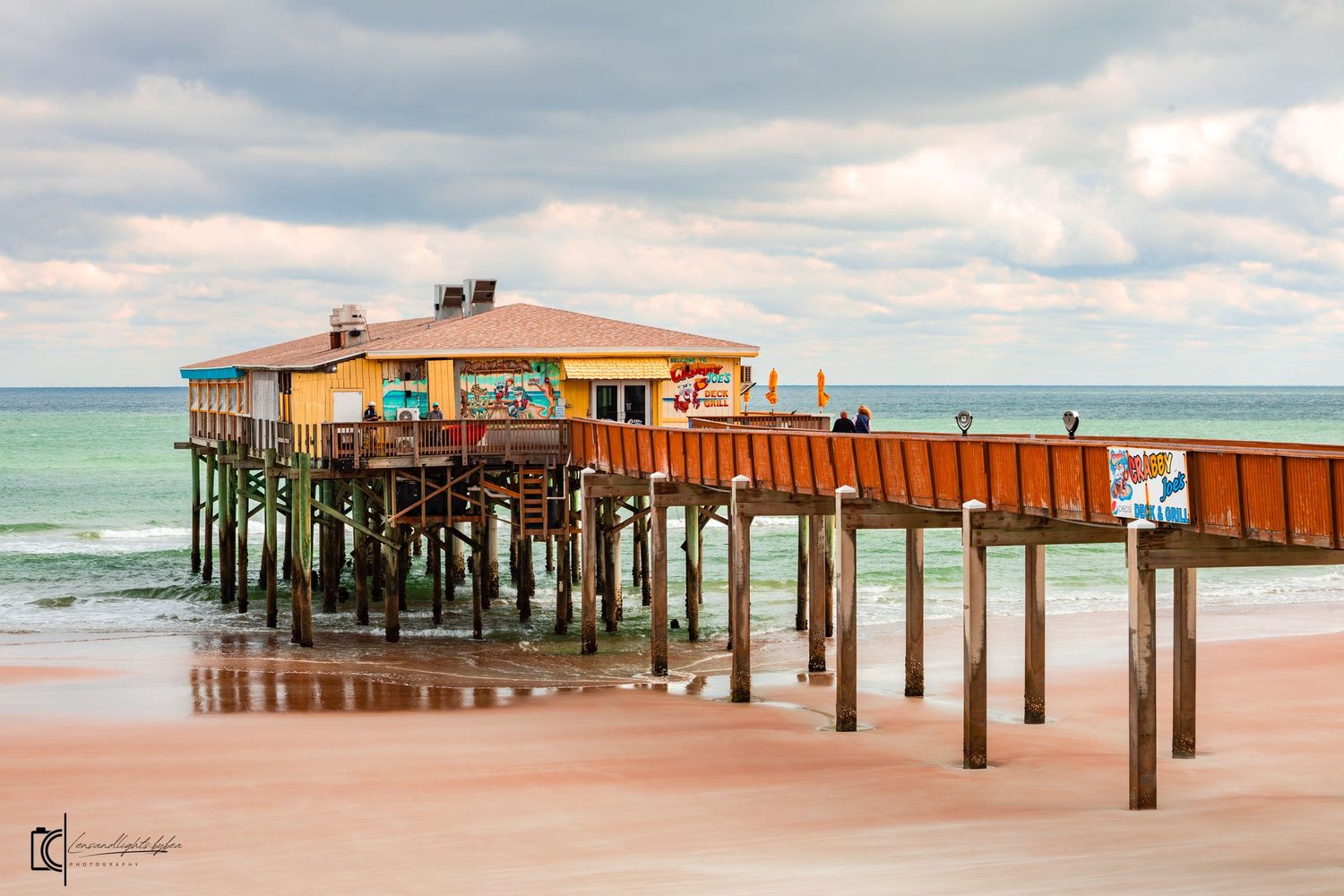 A wooden fishing pier with a rustic restaurant extends over turquoise waters and pink sandy beach under cloudy skies.