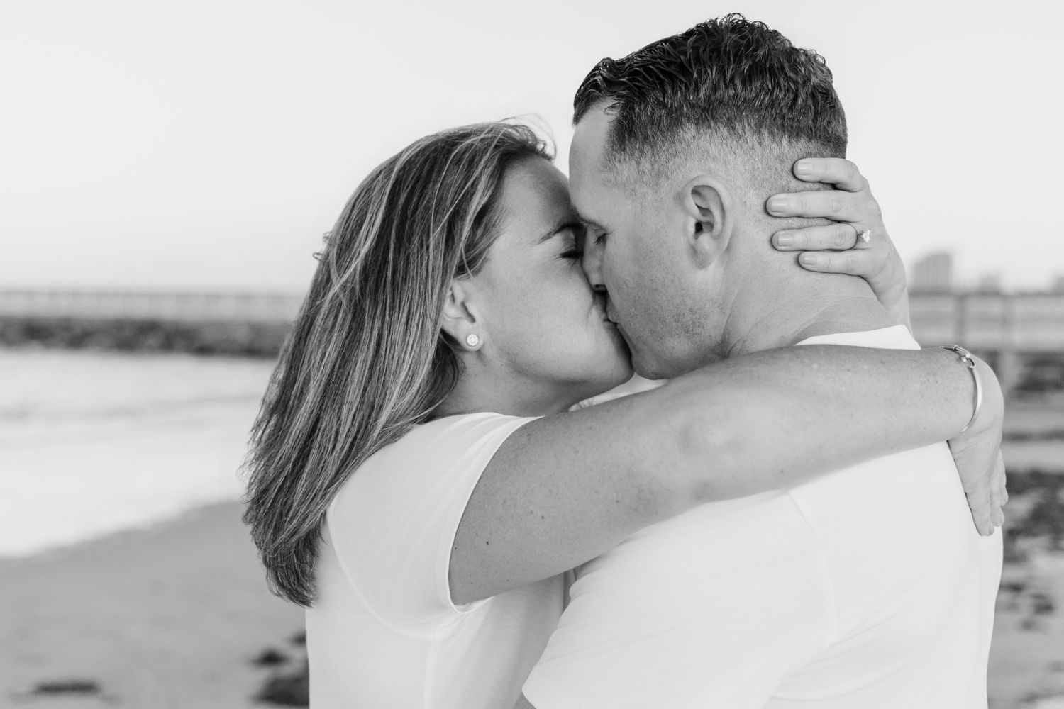 Black and white photo of a couple sharing a romantic kiss on a beach, the woman embracing the man tenderly.