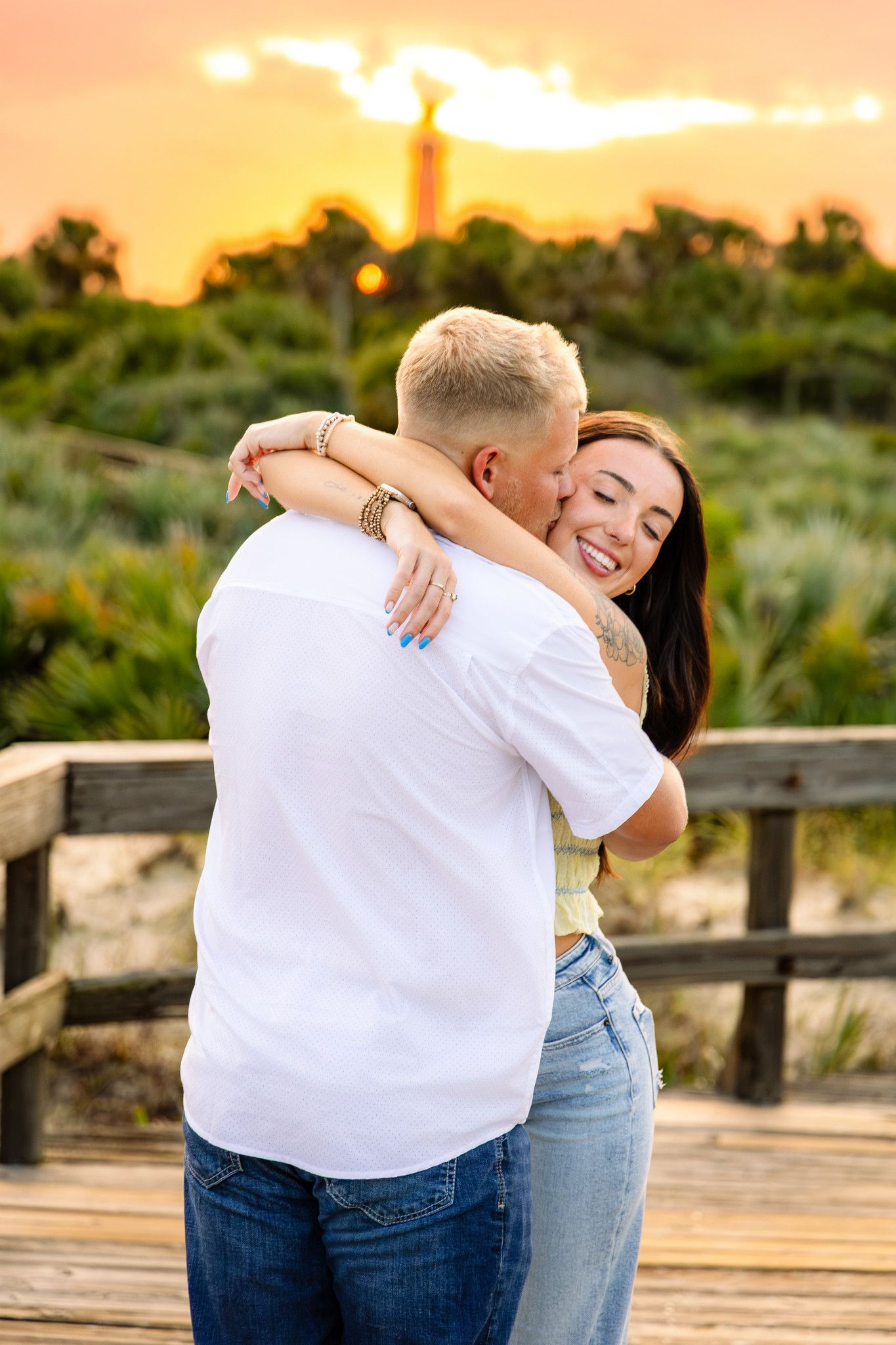 Happy couple hugging on a beach boardwalk at sunset with a golden sky and monument visible behind them.
