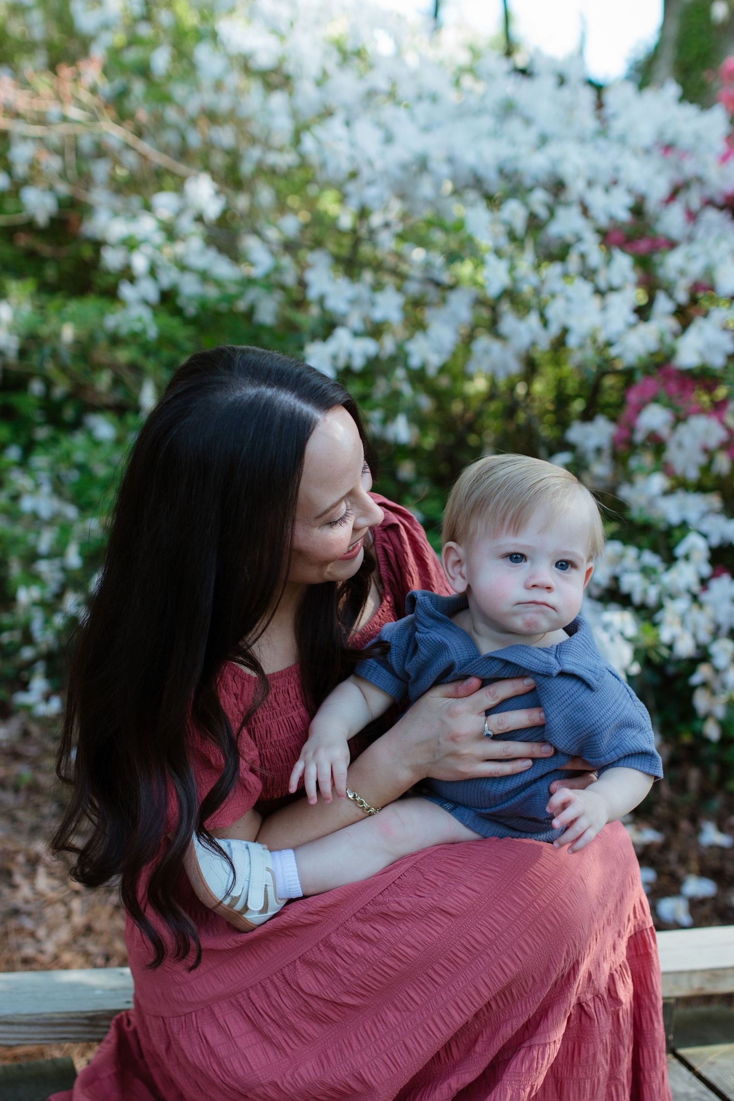 A mother in a  dress holds her  toddler boy outdoors near blooming white and pink flowers at glencairn gardens in rock hill.