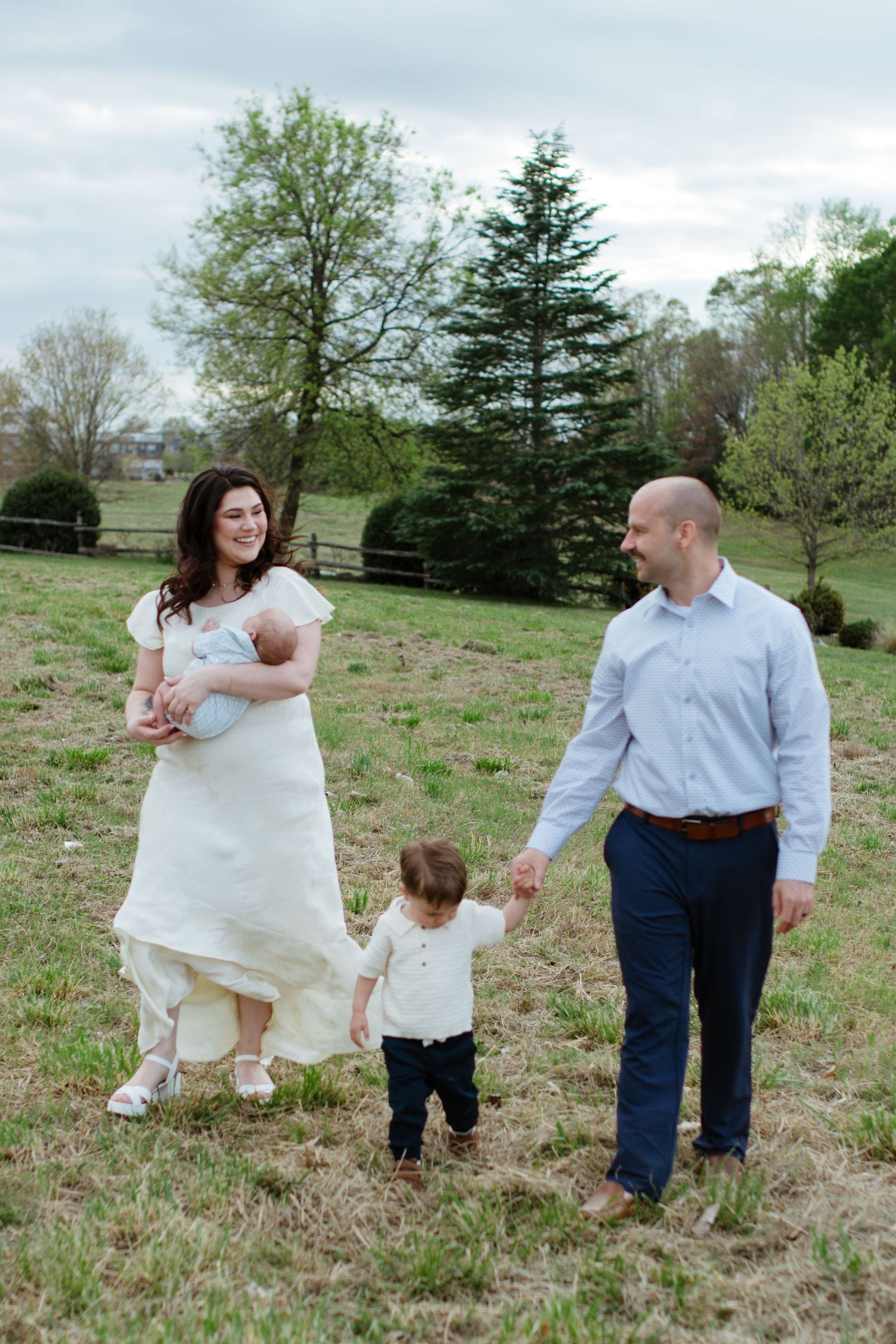 A young family walks outdoors on a green field for a family session, mother holding newborn, father holding toddler's hand.