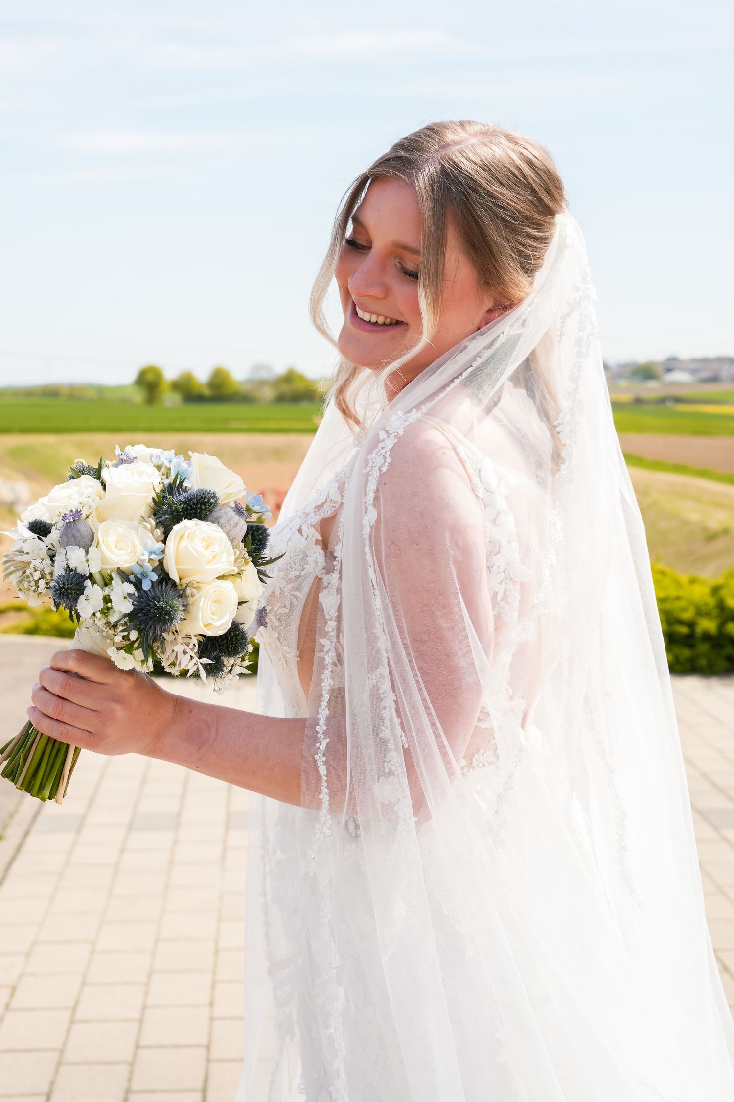 Smiling bride in lace wedding dress and veil holding white and blue floral bouquet outdoors on sunny day.