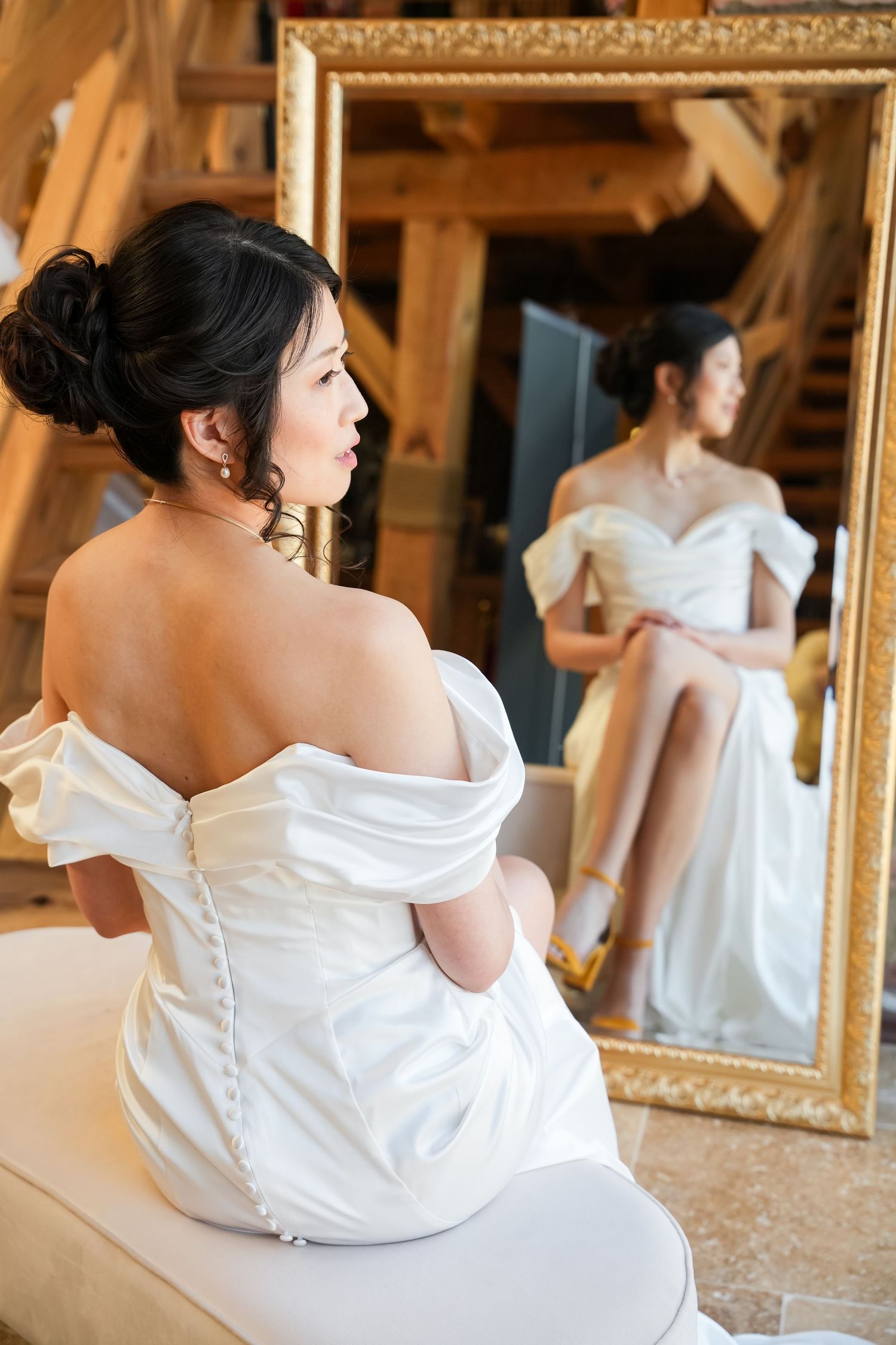 Bride in off-shoulder white satin gown seated before ornate gold mirror, showing button-back detail and elegant updo.