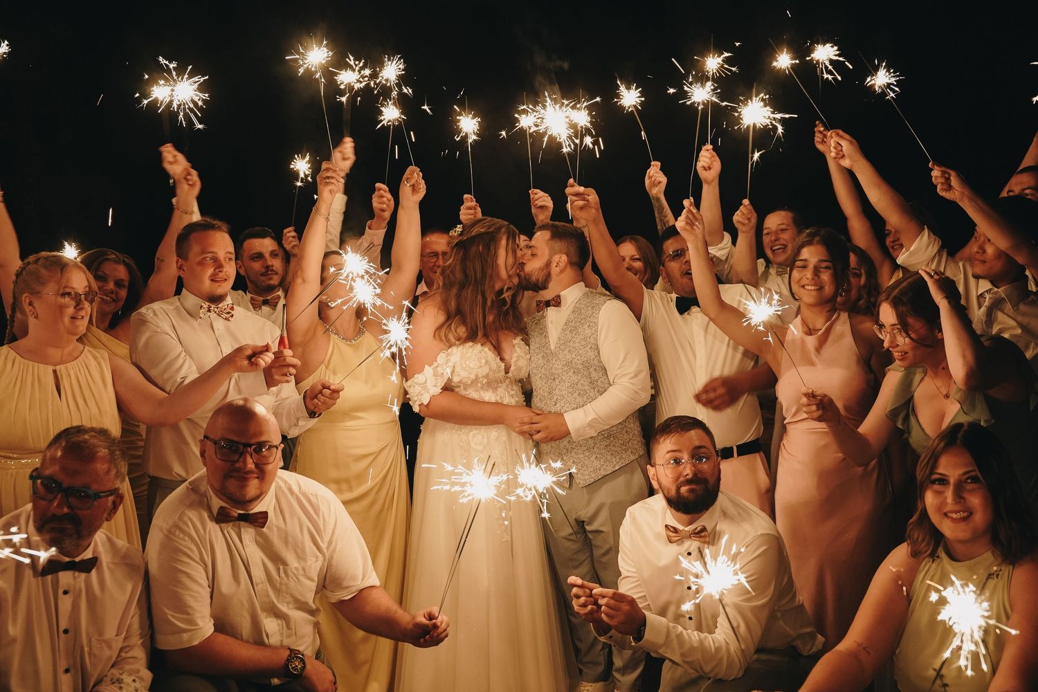 Wedding couple kissing surrounded by guests holding sparklers at night celebration.
