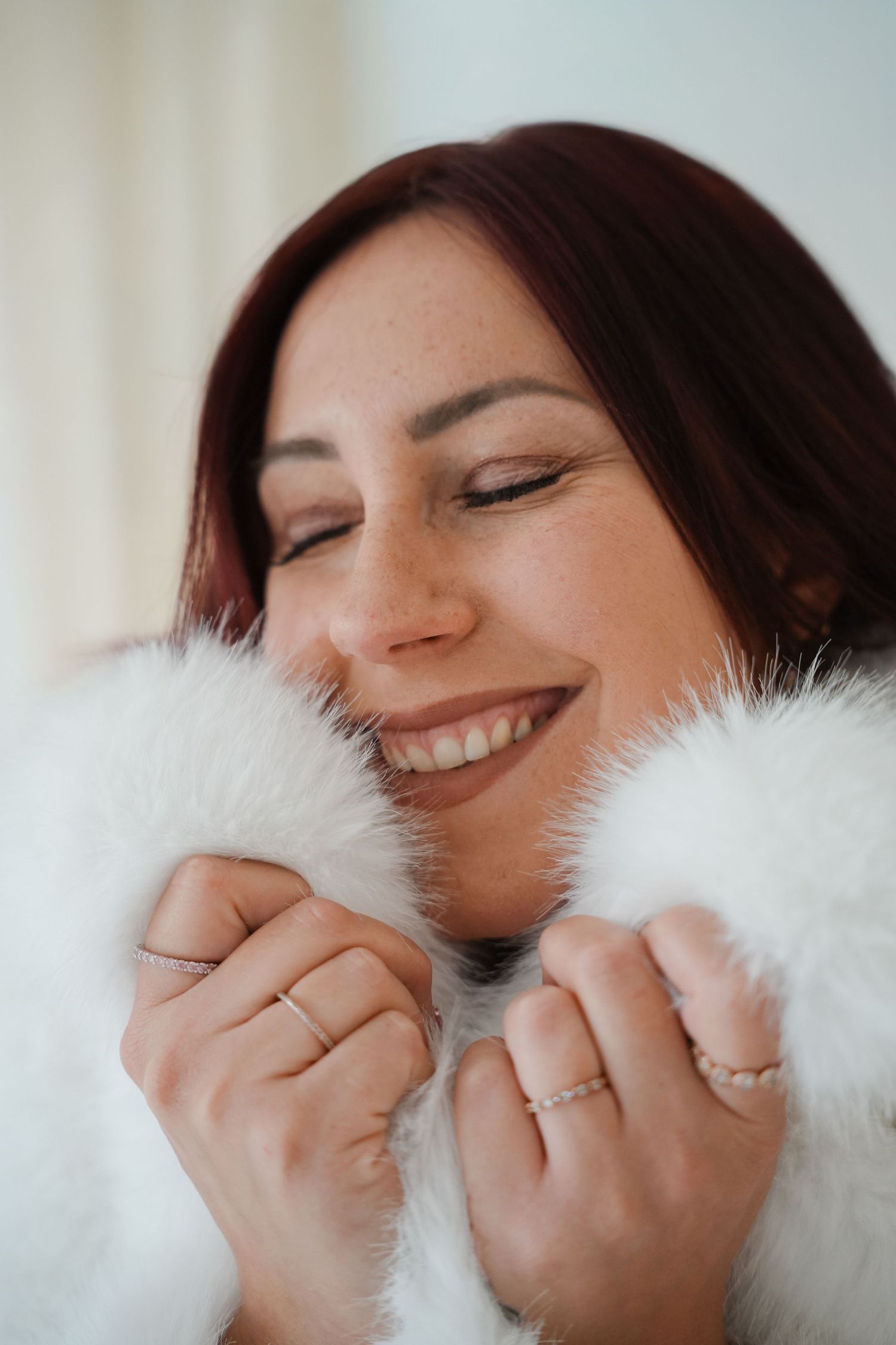 Smiling woman with red hair snuggling into a soft white fur coat with eyes closed, wearing delicate rings.